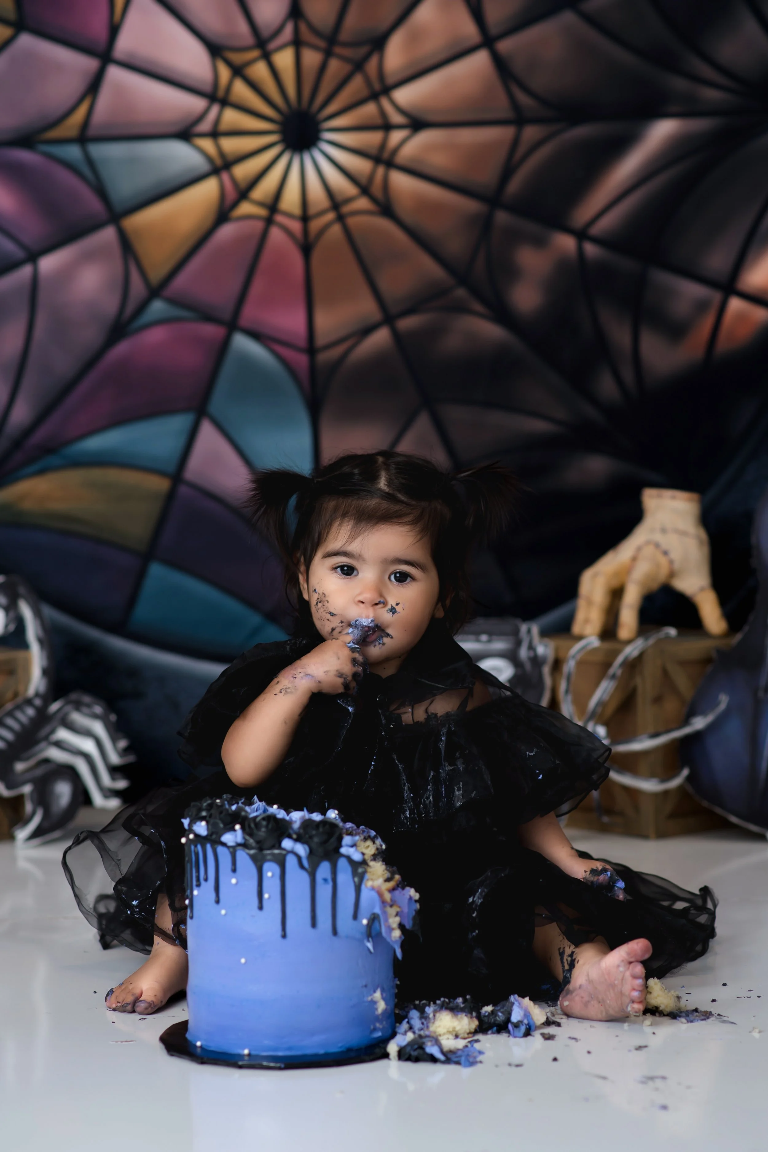 A young girl with dark hair in pigtails, wearing a black dress, sitting on the floor with a partially eaten blue birthday cake. She has cake and frosting on her face, hands, and feet. In the background, there is a large colorful umbrella and some dec
