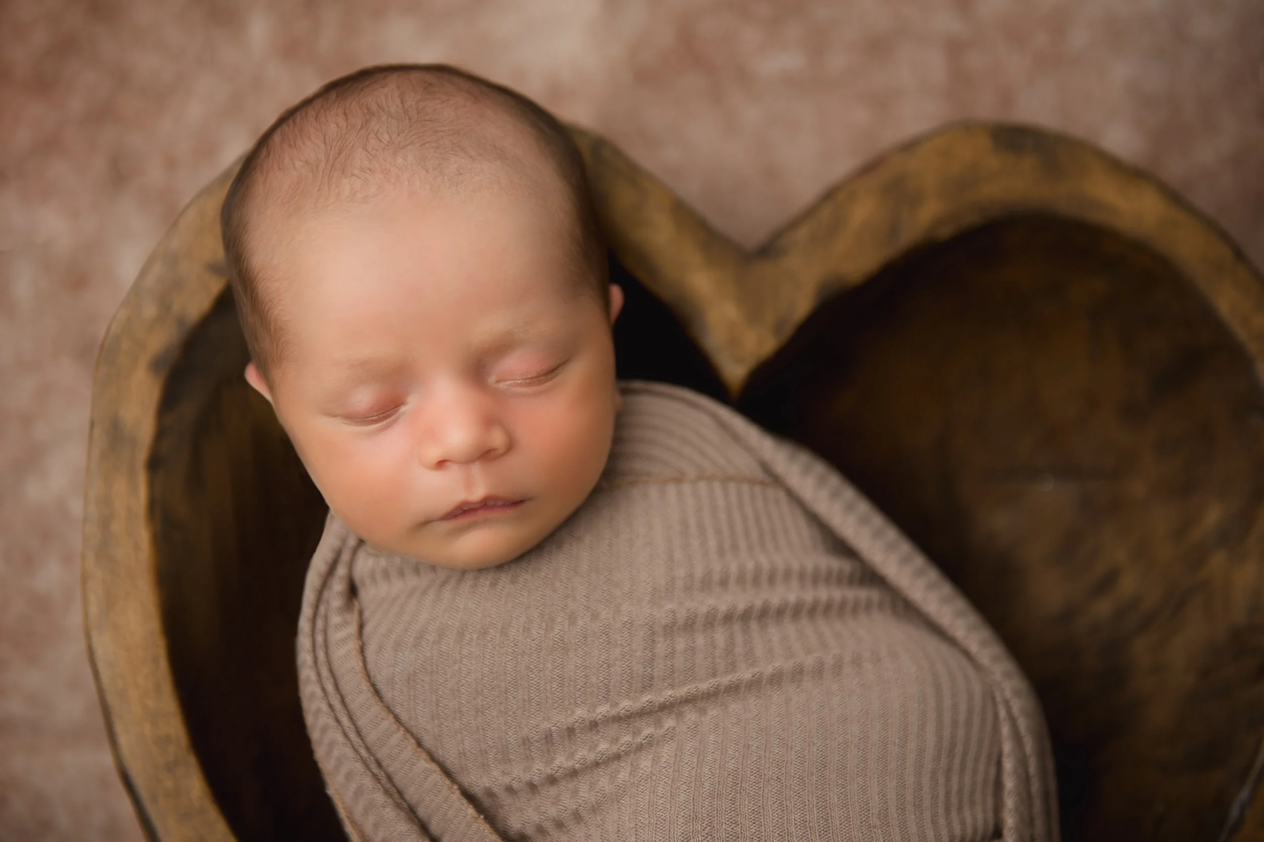 A young baby peacefully sleeping, wrapped in a beige blanket, lying inside a wooden bowl, with a textured beige background.