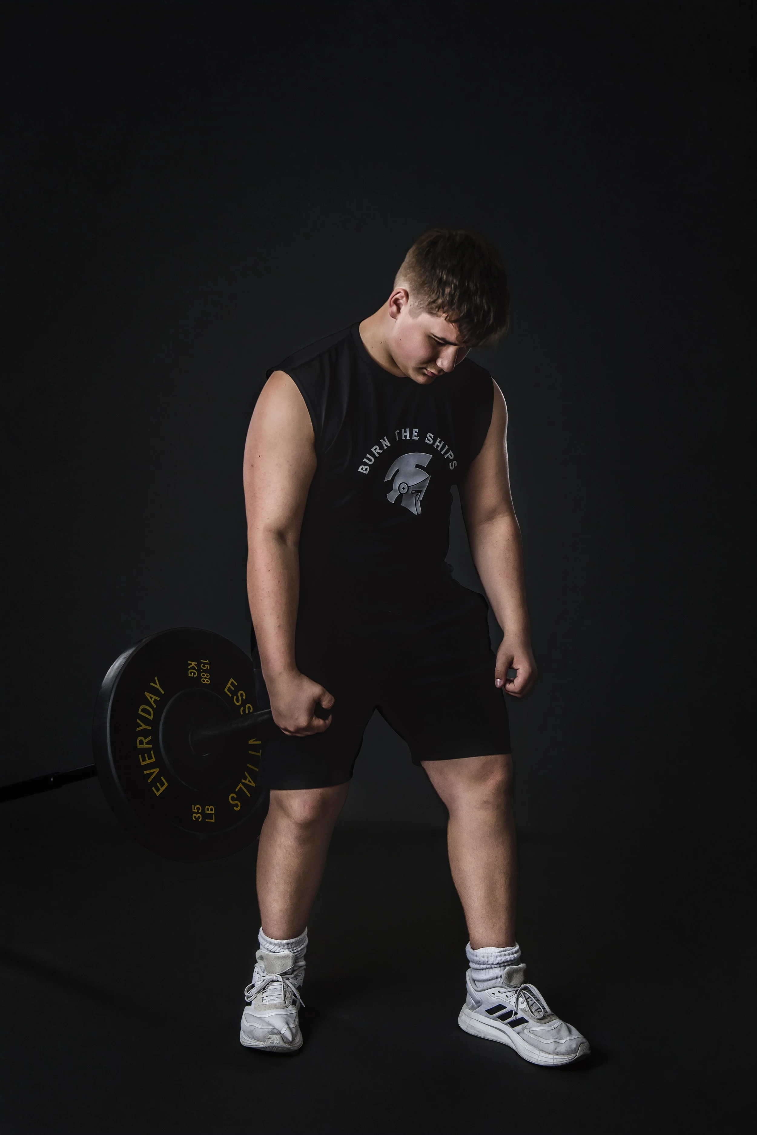 Young man standing in a dark studio holding a barbell with yellow weights, wearing a sleeveless black shirt, black shorts, white sneakers, and socks, with a focused expression.