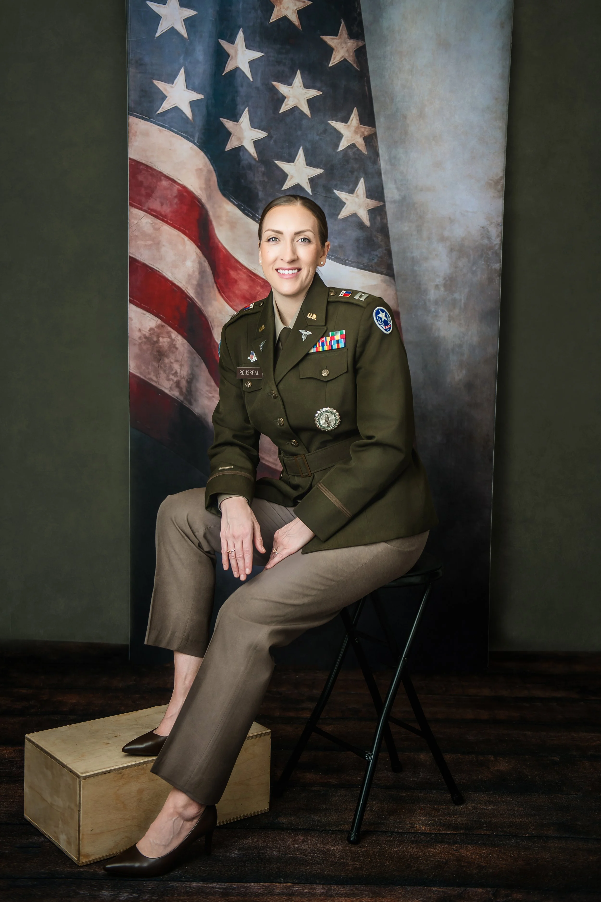 A woman in military uniform sitting on a black folding chair, with one foot on a wooden box, in front of an American flag backdrop.