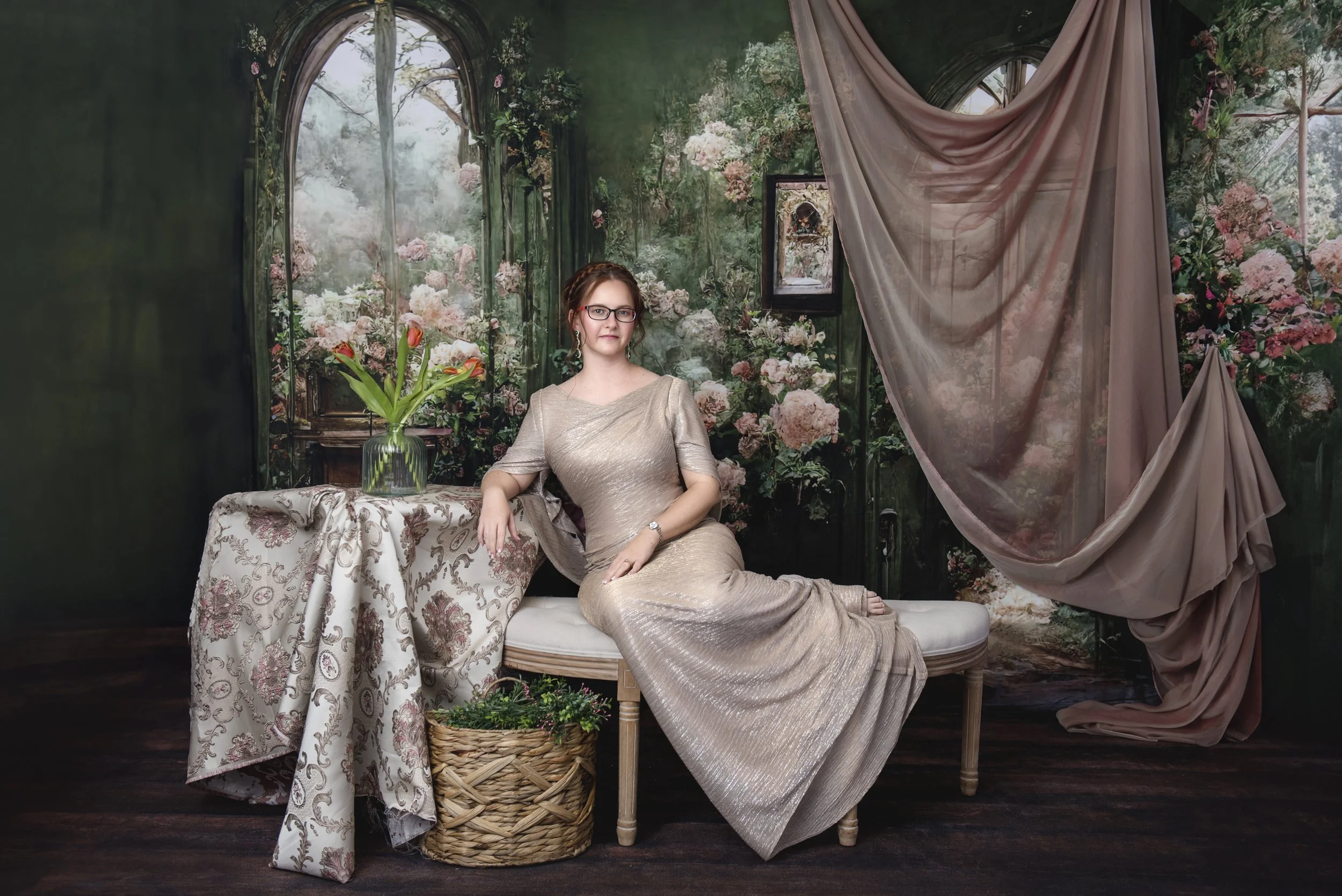 Woman wearing a beige gown sitting on a white bench next to a table with a green vase of pink and red tulips, in front of a backdrop of pink and white flowers with large arches and draped fabric.