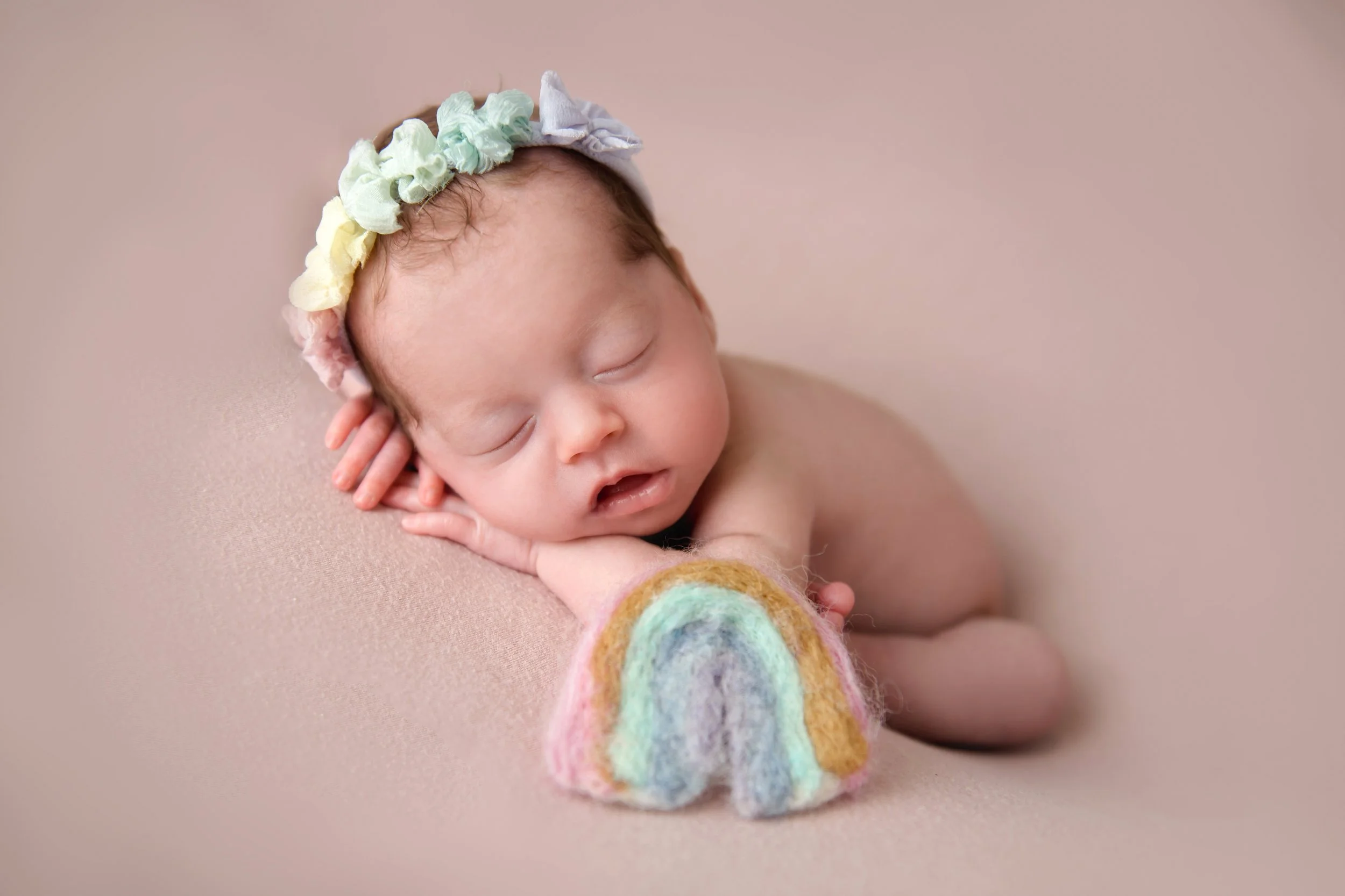 Fine art newborn portrait of a sleeping baby wearing a colorful floral headband, posed beside a felt rainbow on a soft textured surface.
