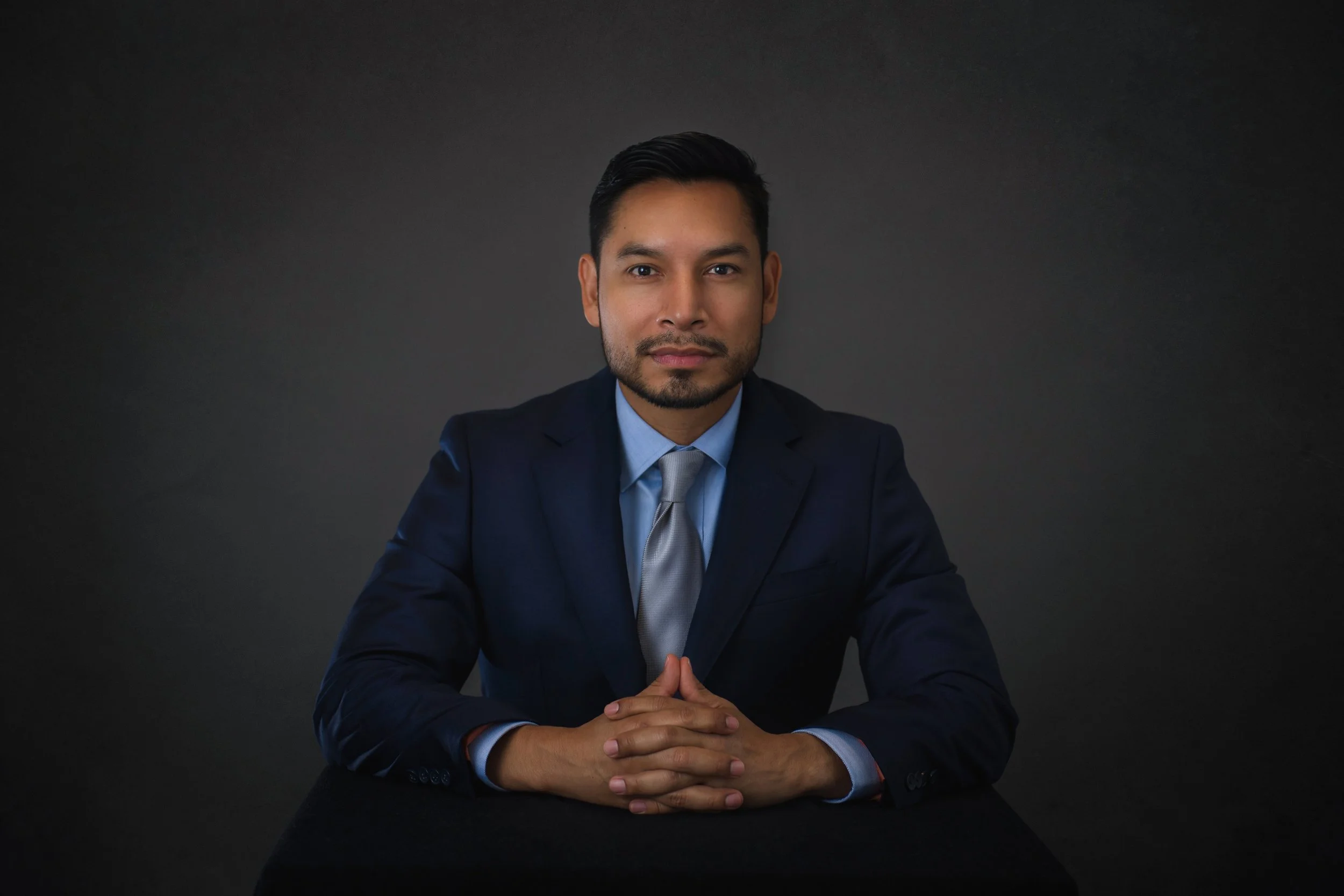 Portrait of a man in a navy suit, light blue shirt, and grey tie, seated at a black table against a dark background, with hands clasped.