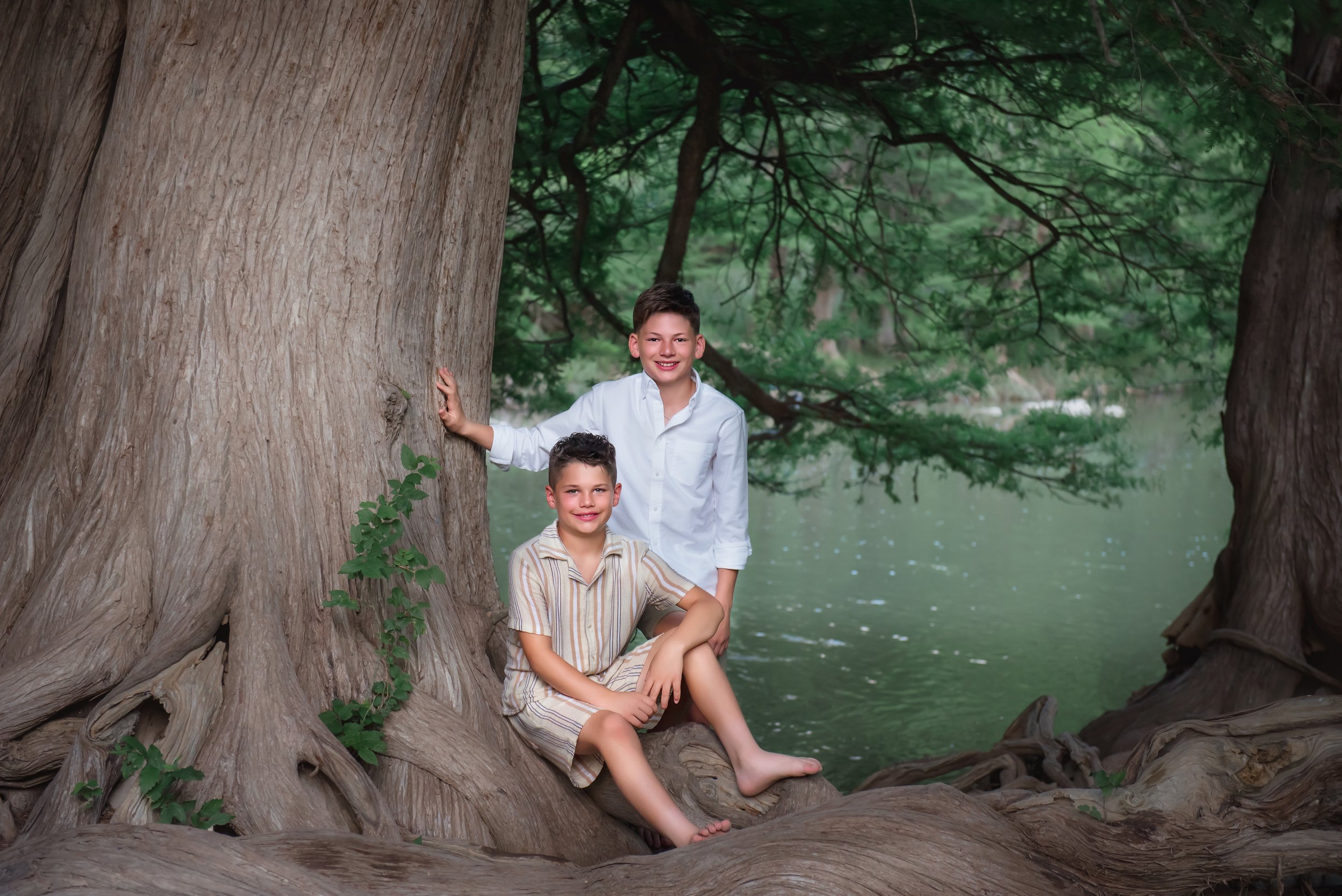 Two boys sitting and standing on large twisted roots of a massive tree by a body of water, surrounded by lush green branches and leaves in the Hill Country.