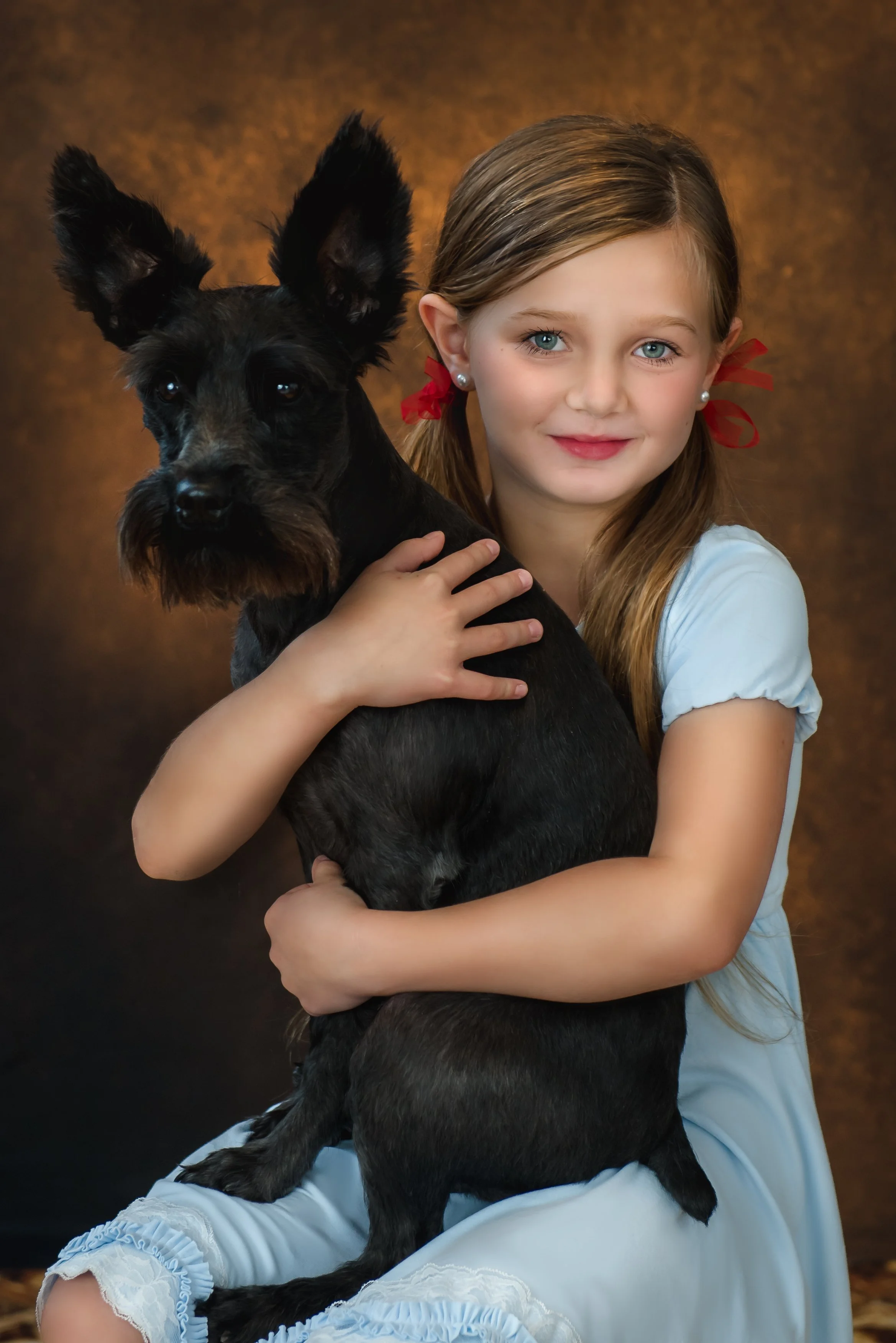 A young girl with blue eyes, light brown hair, and red ribbons in her hair, holding a small black dog with upright ears and a beard, against a brown textured background.