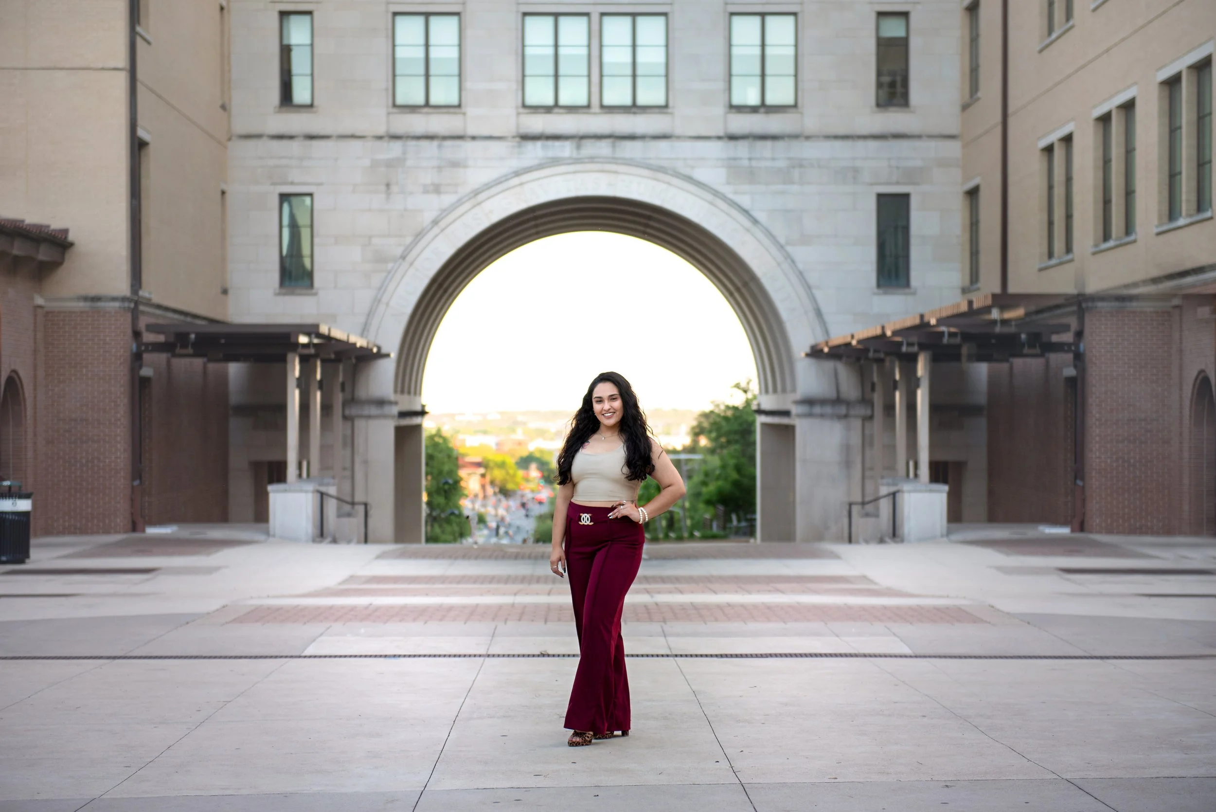 A woman with long dark hair wearing a beige sleeveless top, maroon high-waisted pants with a buckle, and heels, standing confidently with one hand on her hip in front of a large stone archway in an urban setting.