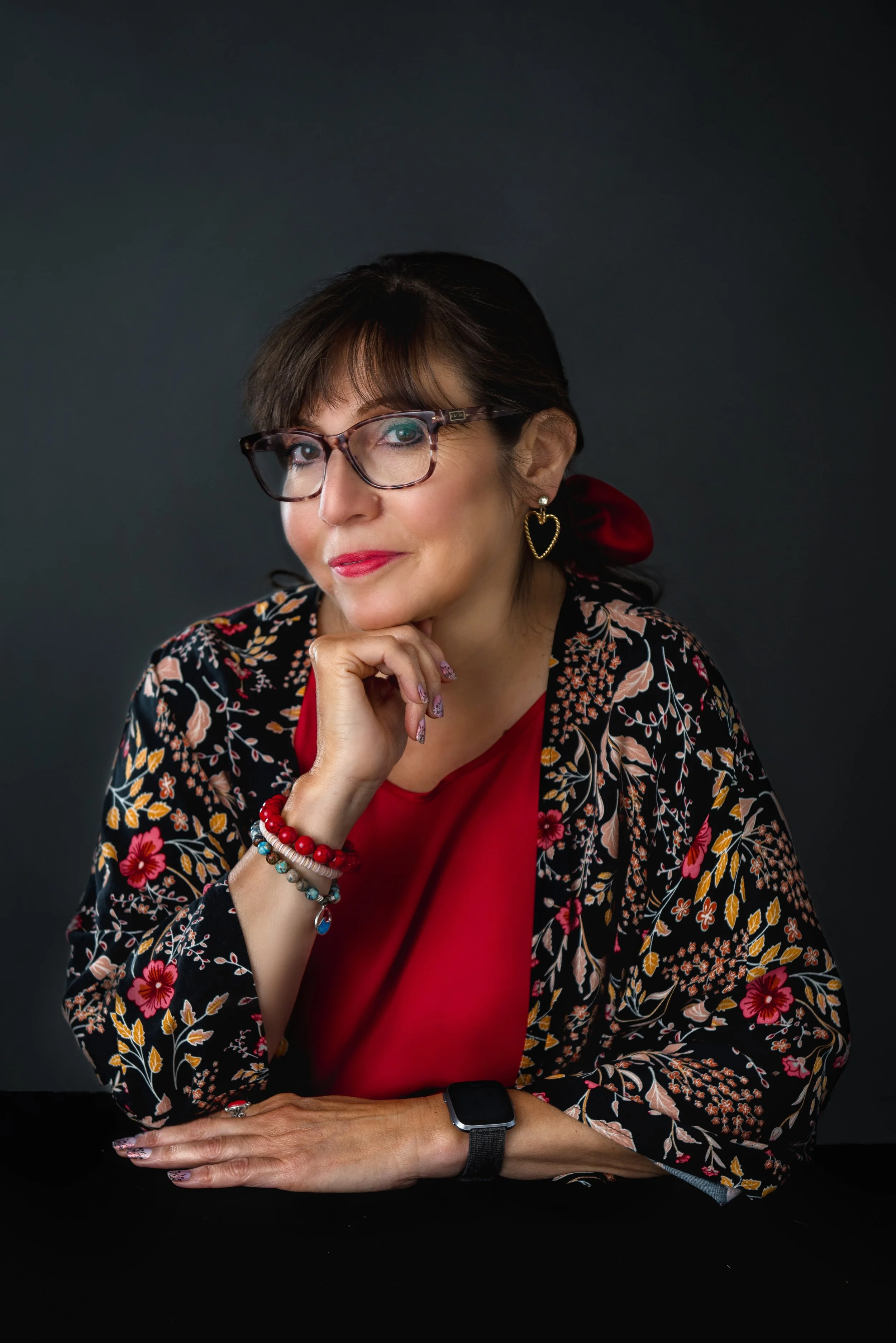 A woman wearing glasses, a red blouse, and a floral blazer, with jewelry including heart earrings and beaded bracelets, sitting at a black table against a dark background in our Spring Branch studio.