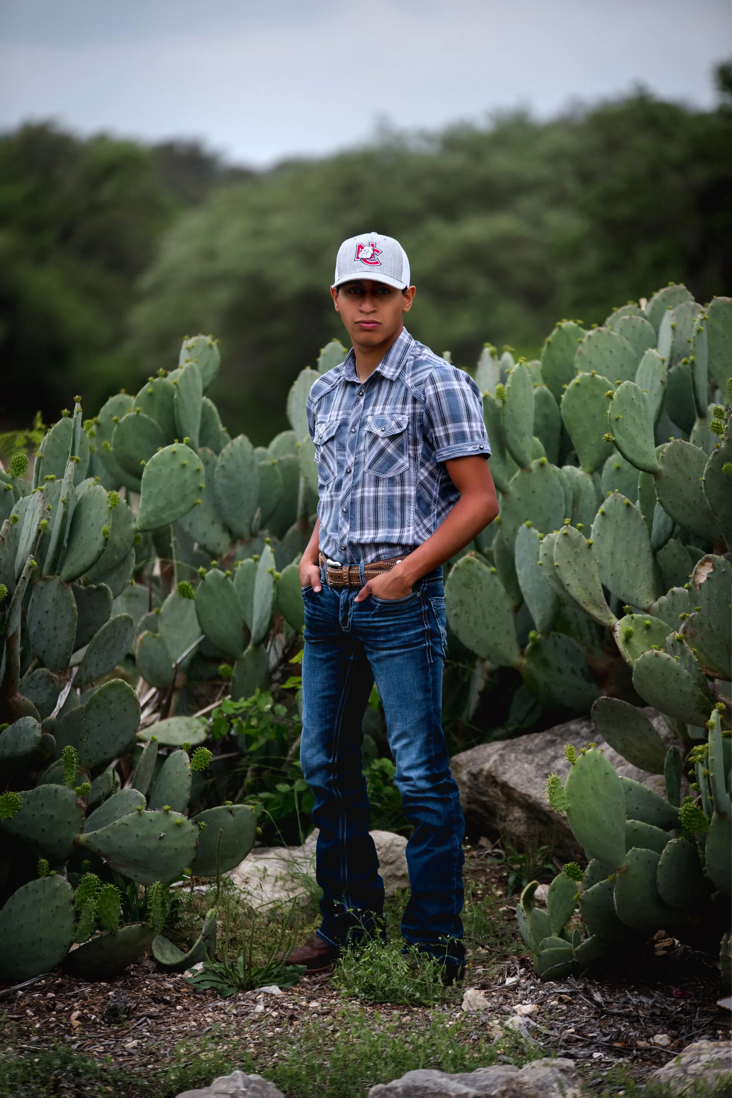A young man in a plaid shirt and jeans standing amidst large prickly pear cacti with a blurred green landscape background.