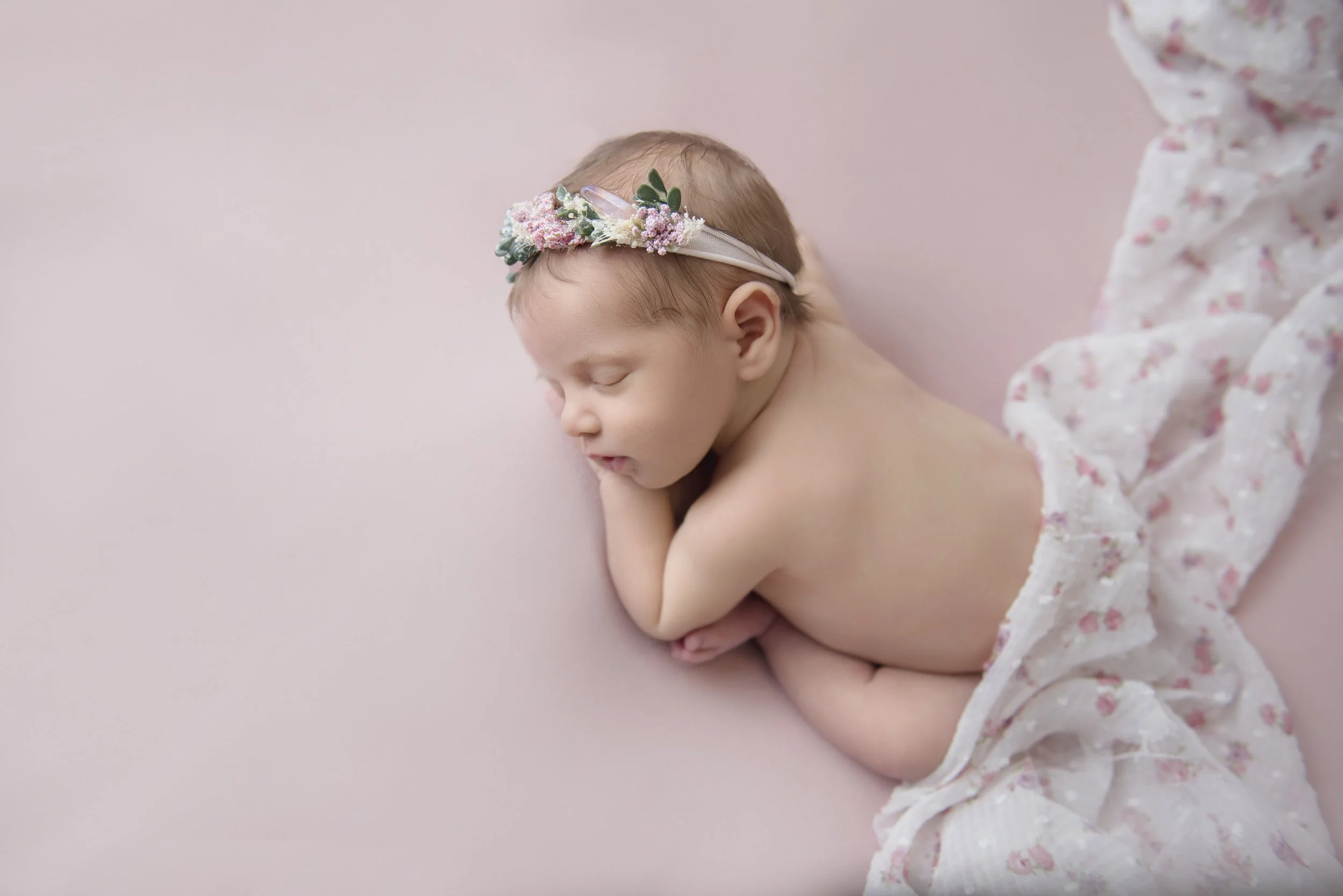 A sleeping baby girl on a pink background, wearing a floral headband and a lacy blanket.