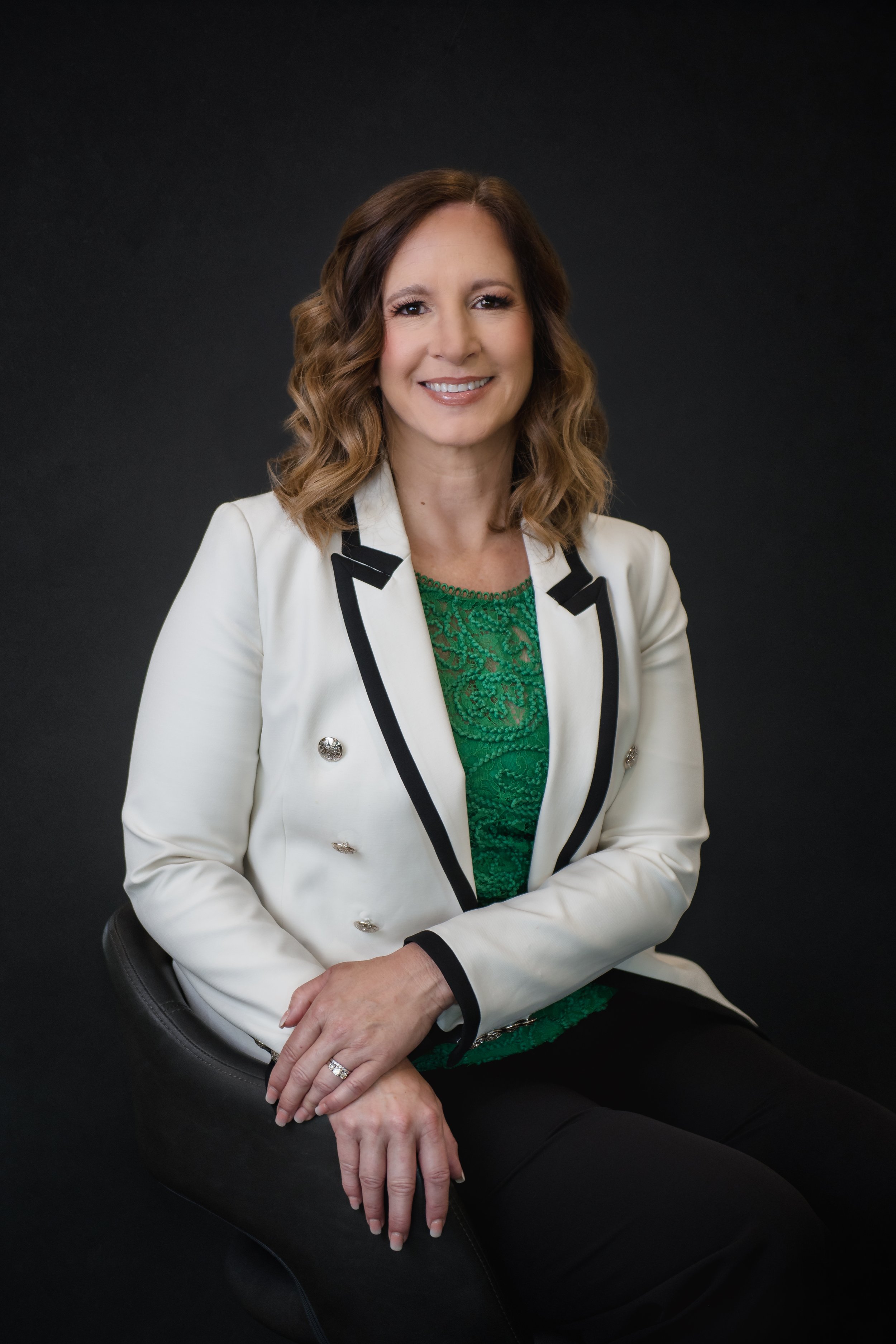 A woman with wavy brown hair, wearing a white blazer with black trim and gold buttons, a green lace top, and black pants, sitting on a black chair against a dark background, smiling.