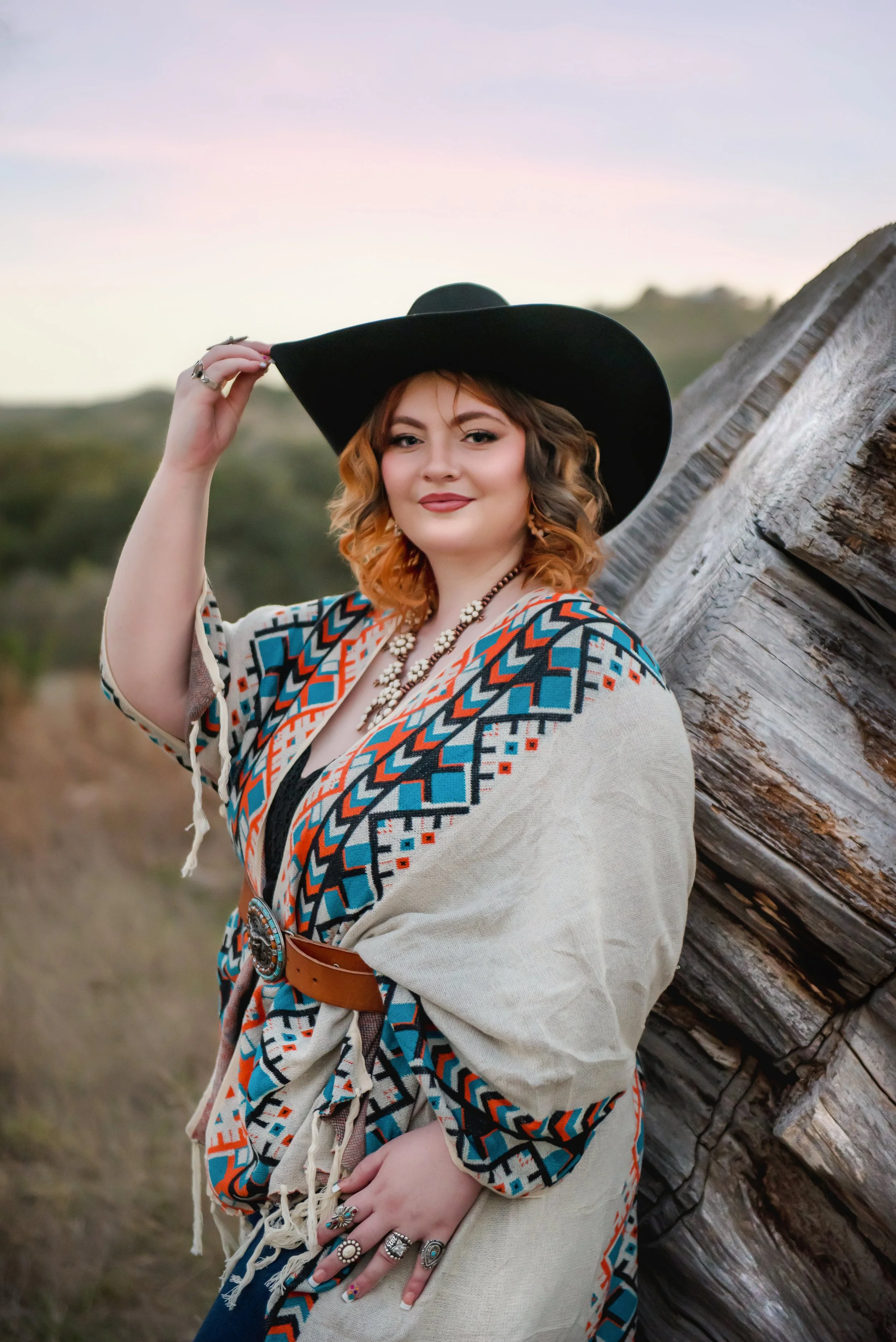 A woman with curly hair in shades of blonde and brown, wearing a large black hat, tribal-patterned shawl, and a chunky necklace, standing outdoors next to a large tree with a blurred natural landscape background.