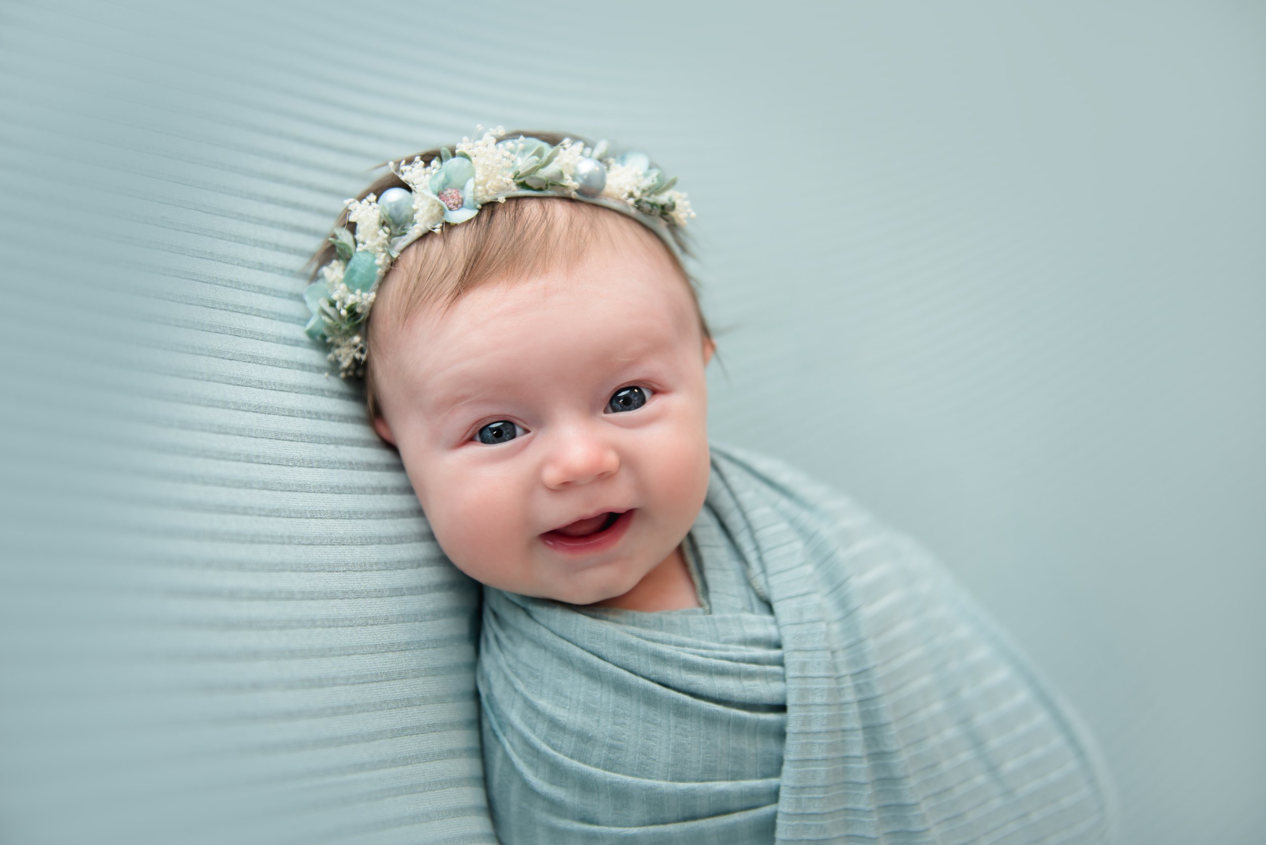 A happy baby with blue eyes wearing a floral headband and a light blue striped swaddle blanket, lying on a light blue textured surface.