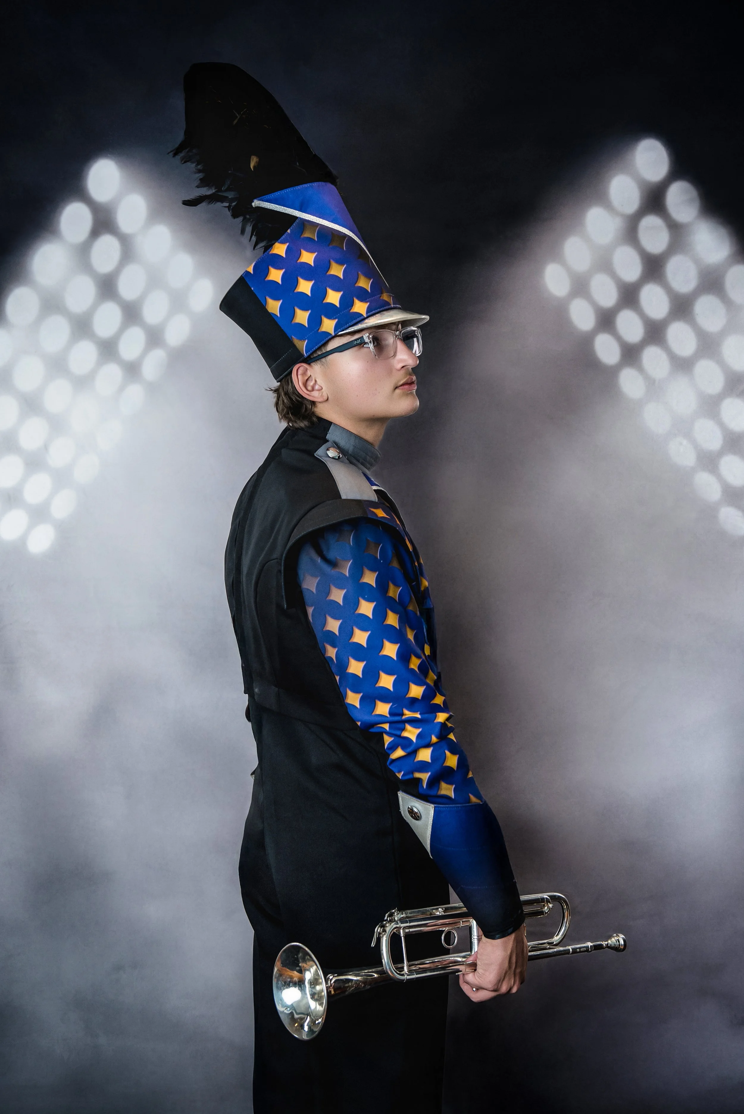 A young man dressed in a marching band uniform with a blue and yellow patterned hat and jacket, holding a trumpet, standing against a dark background with blurred white circular lights.