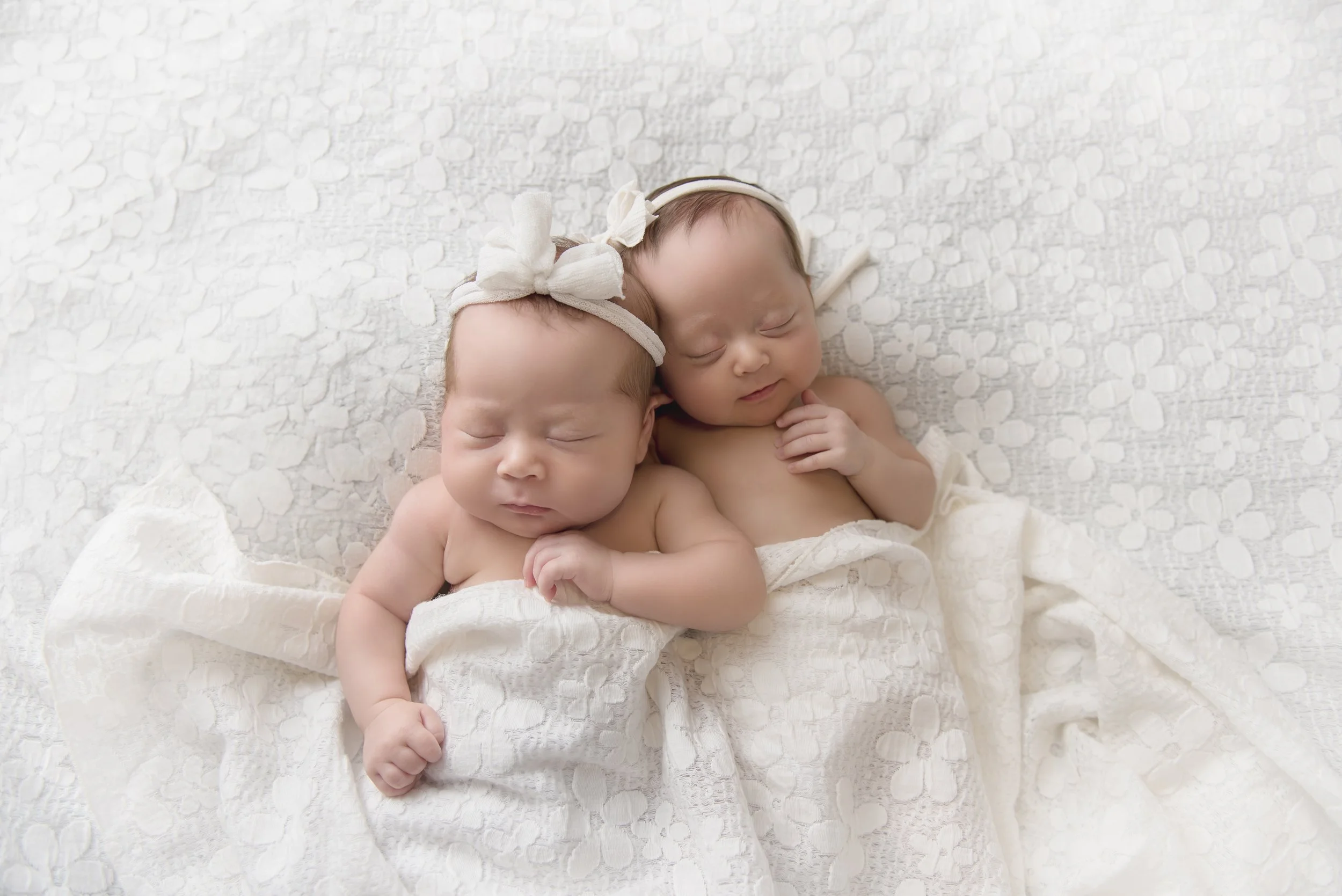Two sleeping babies wrapped in a white blanket, with one wearing a headband with a bow.