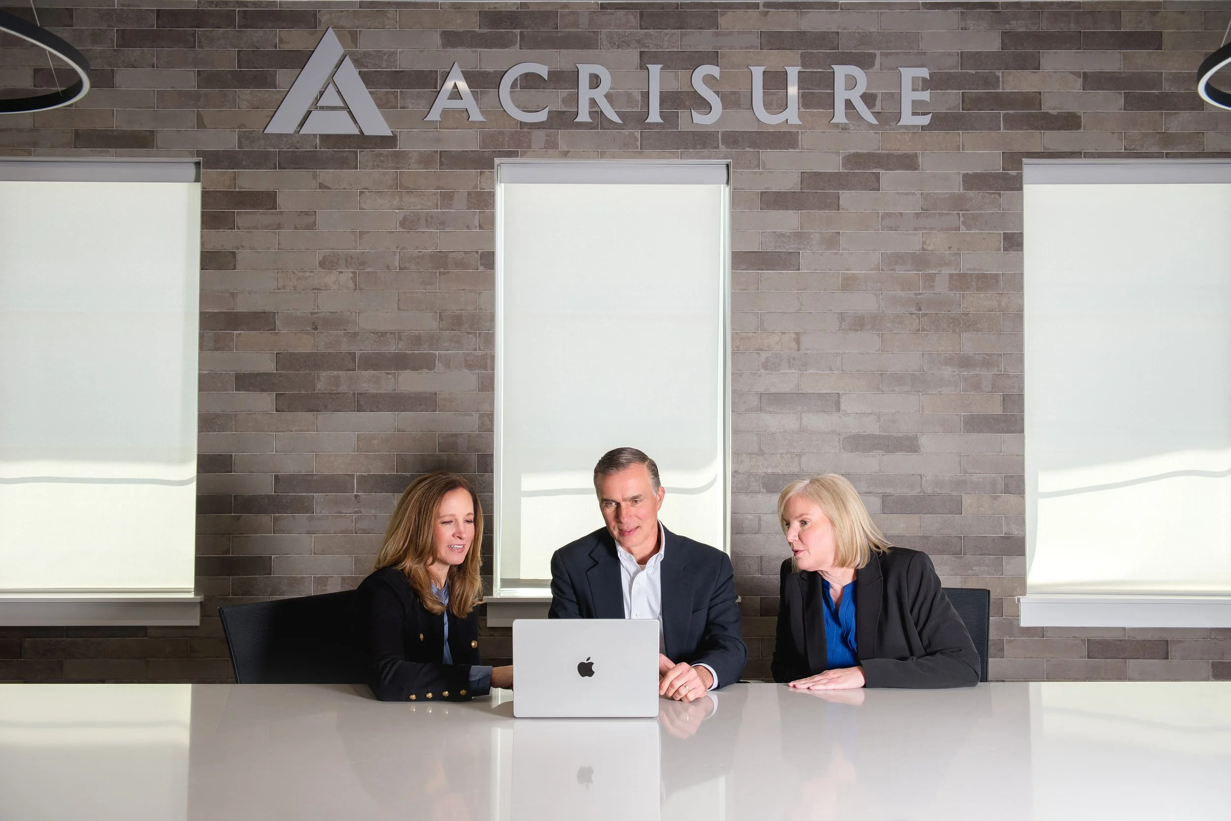Three business professionals, two women and one man, sitting at a white conference table with a laptop open in front of them, engaged in a discussion in a modern office with brick walls and large windows.
