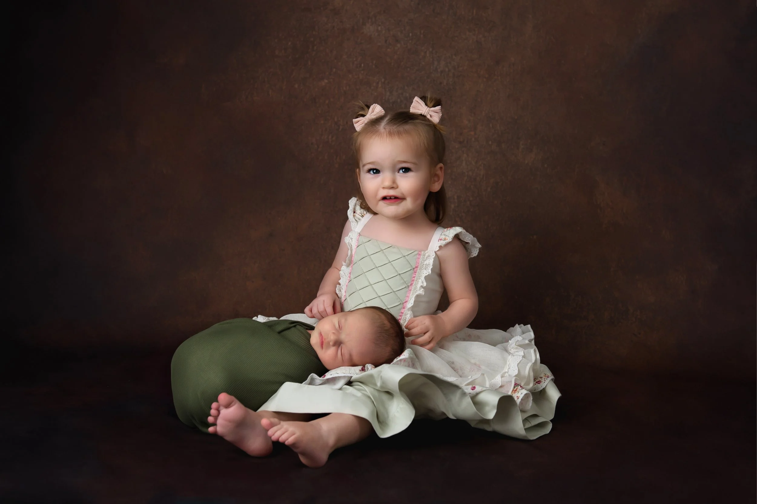 A young girl with two pink bows in her hair sits with a sleeping baby on her lap in front of a dark brown background.