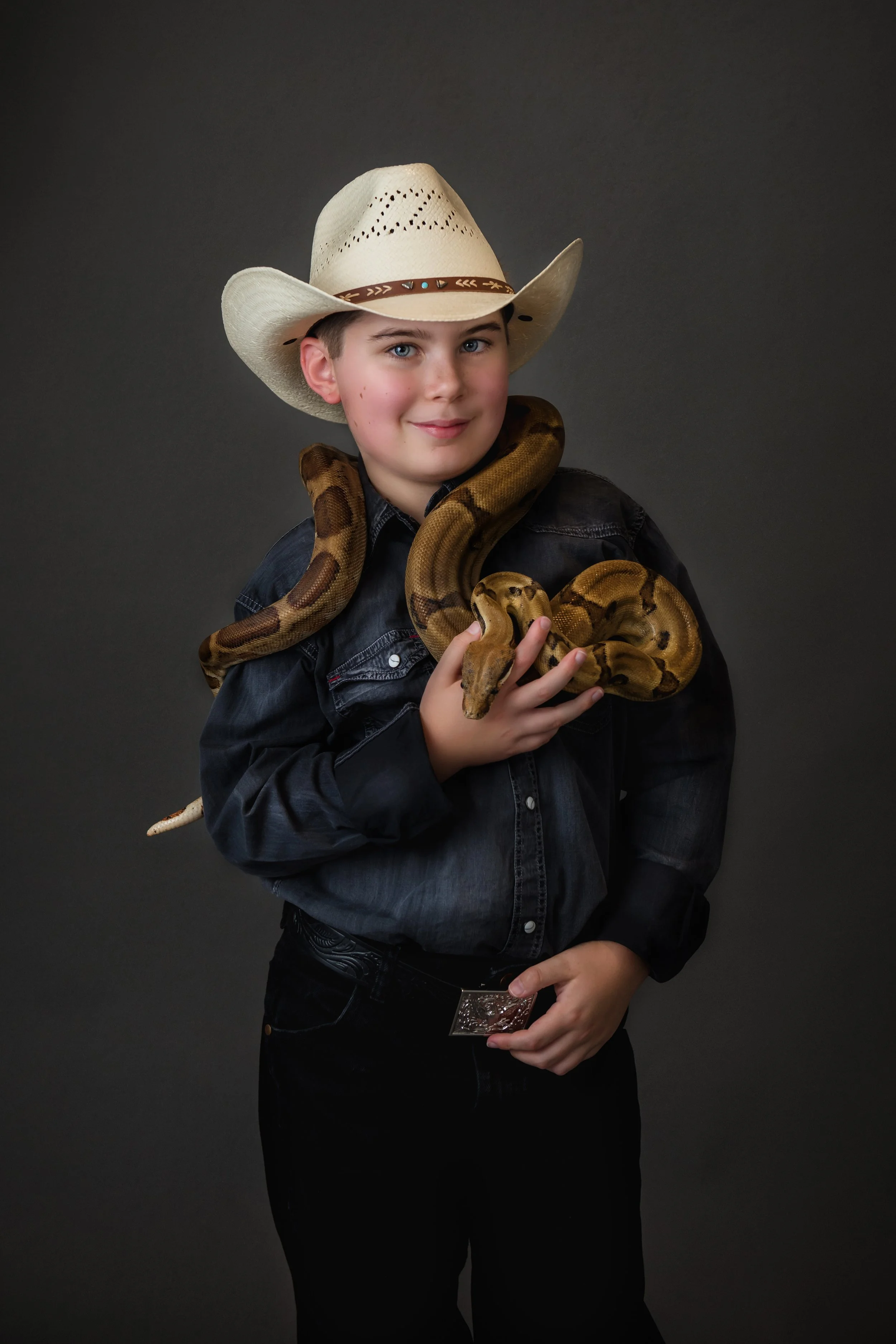 In our Spring Branch studio a young boy wearing a cowboy hat and a black denim shirt is holding a snake around his neck and arm against a dark background.