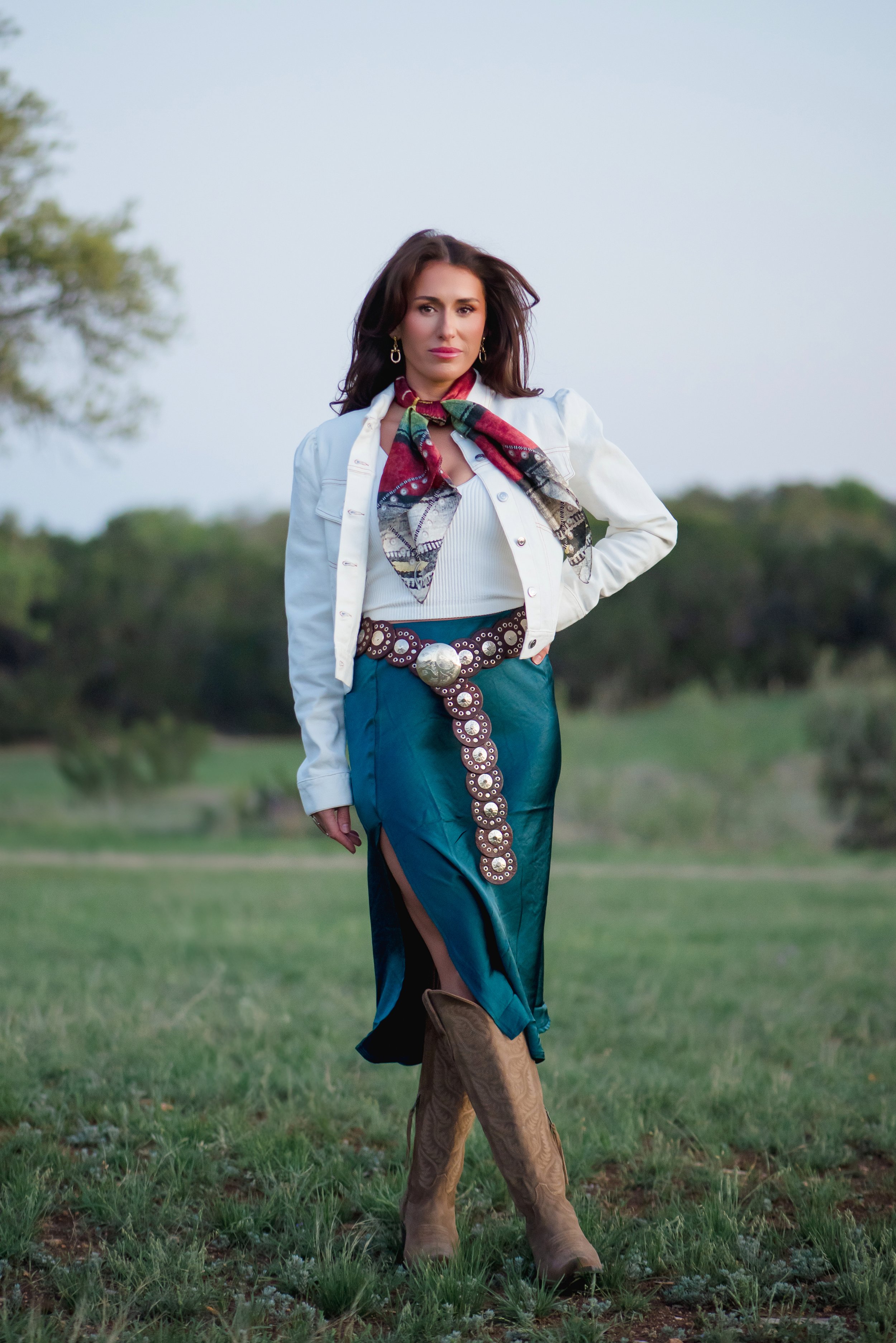 A woman standing outdoors on a grassy field, wearing cowboy boots, a teal skirt with a slit, a white jacket, and a colorful scarf around her neck.