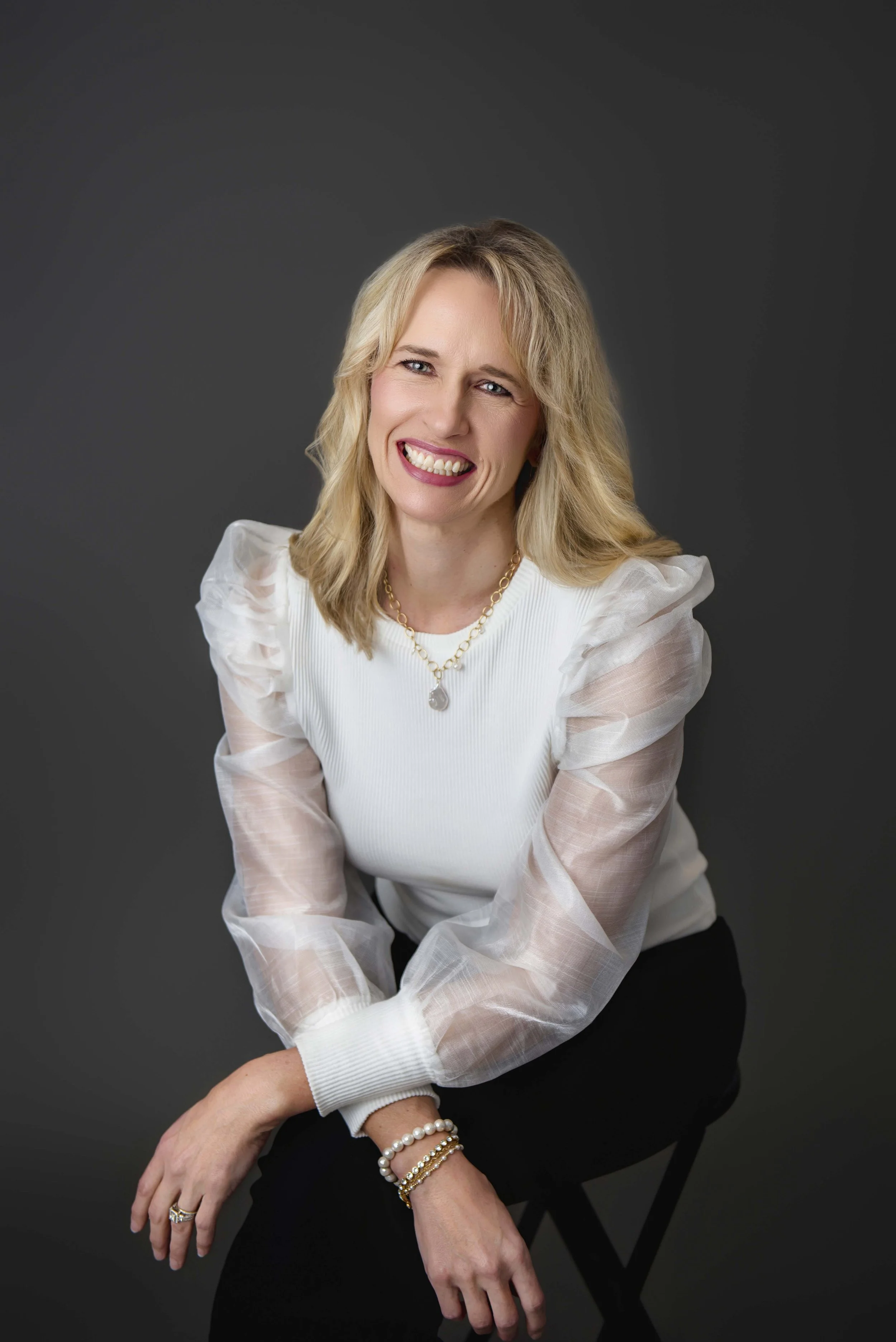 Portrait of a blonde woman wearing a white blouse with sheer puffed sleeves, smiling, sitting against a dark background.