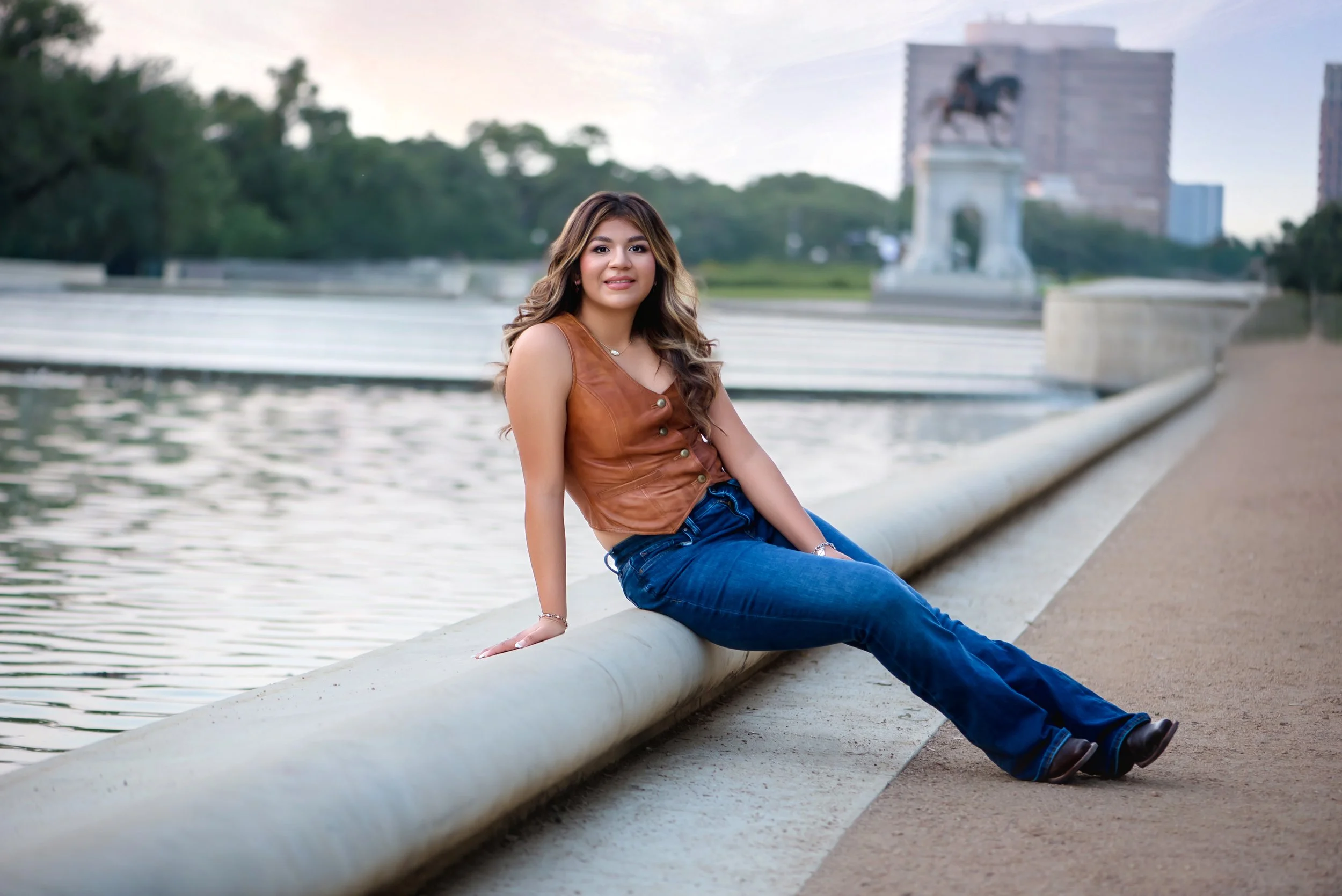 A young woman with long hair sitting on a concrete edge by a body of water, wearing a brown sleeveless top and blue jeans, with a cityscape and a monument in the background.