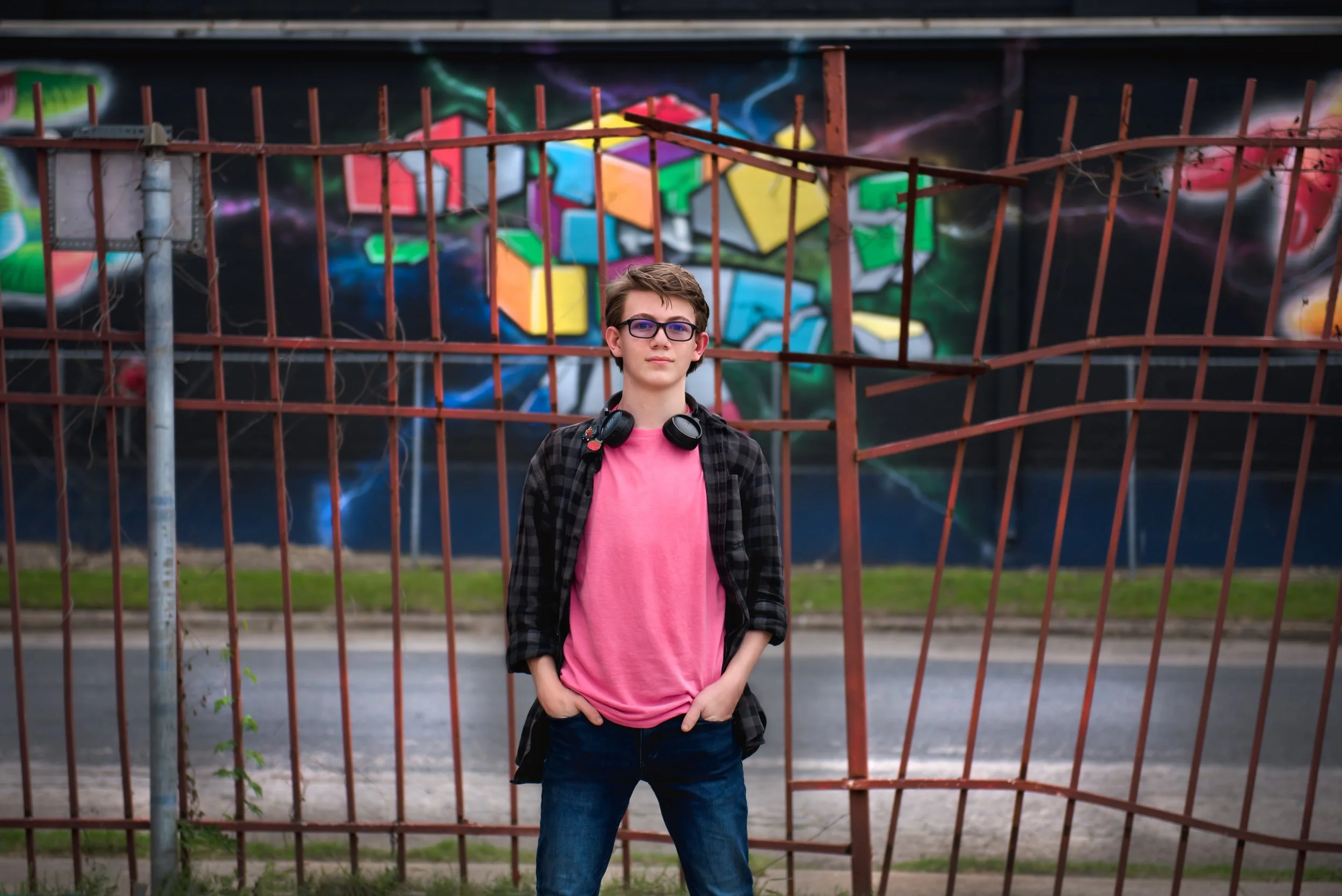A young man wearing glasses, a pink t-shirt, and a black plaid shirt standing with his hands in his pockets in front of a rusted metal fence and a colorful graffiti mural.