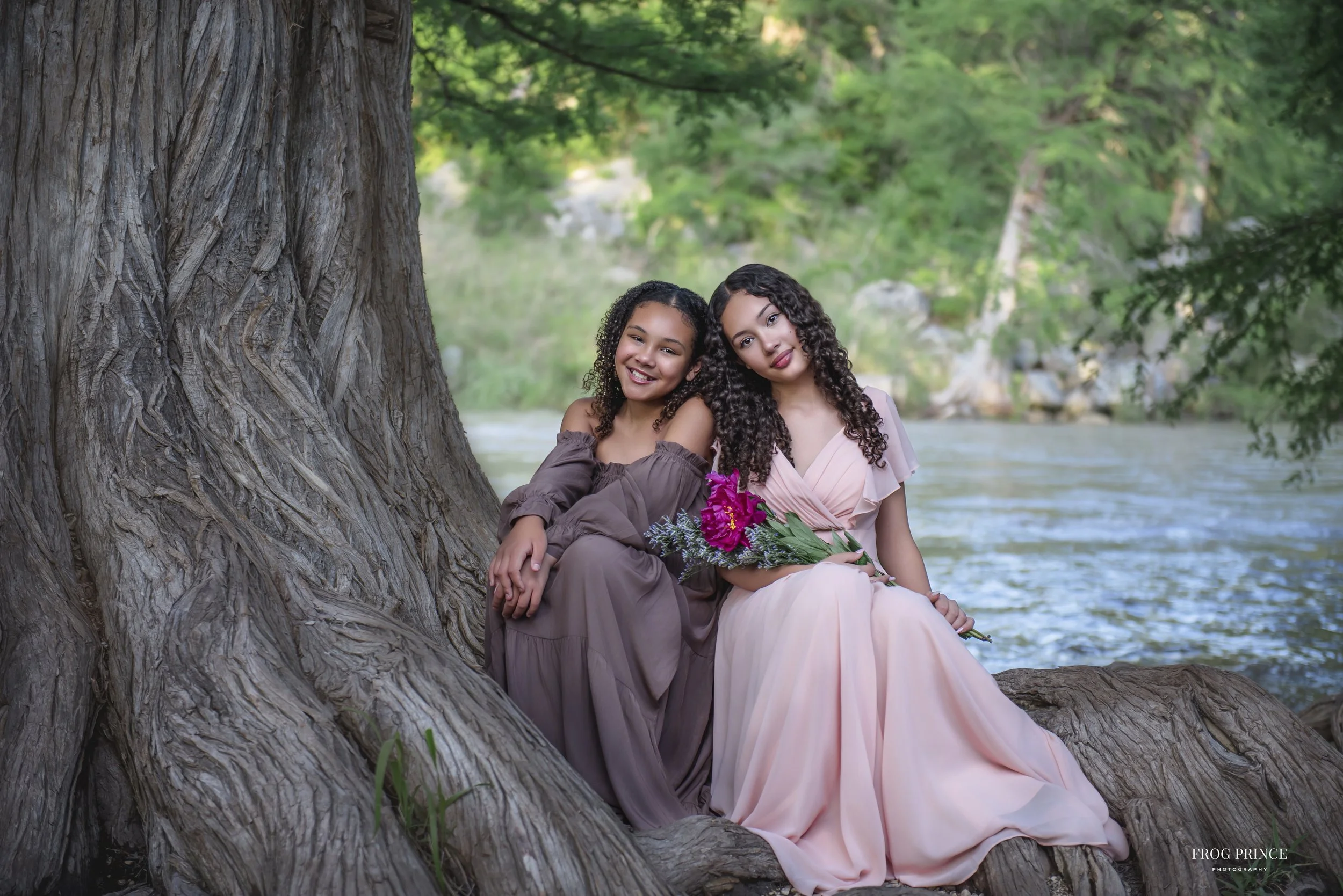 Two young women sitting against a large tree trunk by a river, smiling and holding flowers, surrounded by lush green trees.