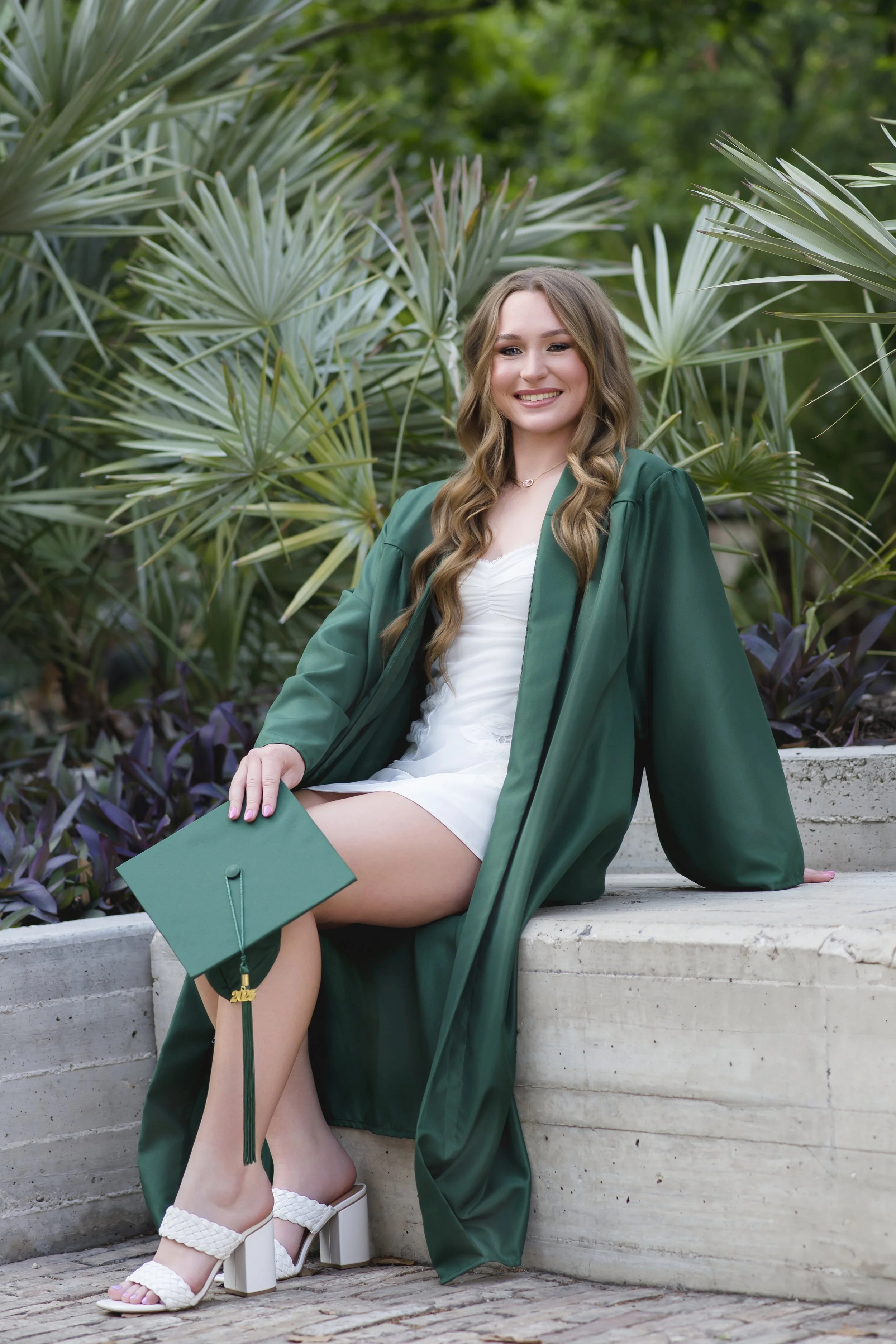 A young woman in a white dress and green graduation gown sits outdoors on a concrete ledge, holding a green mortarboard, with lush green plants in the background.