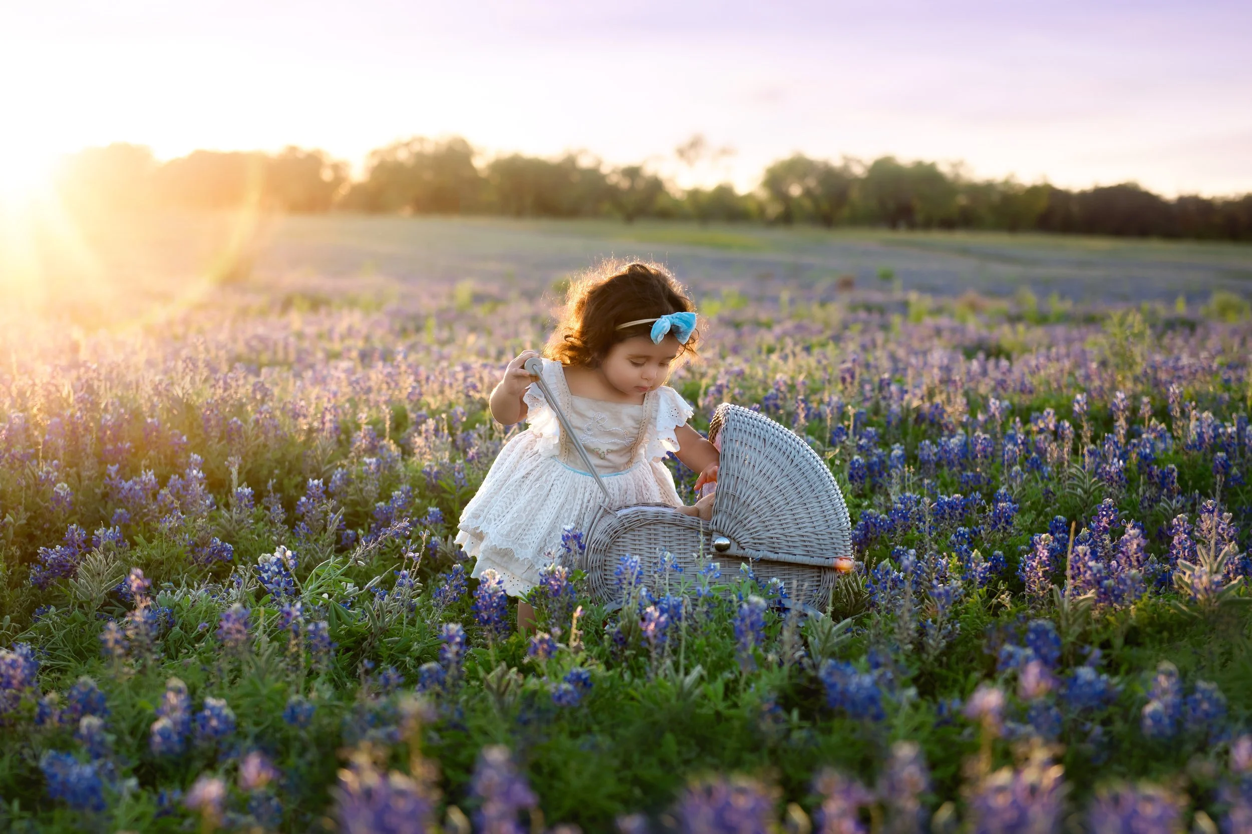 Young girl in a white dress with a blue bow in her hair, standing in a field of purple and blue flowers at sunset, with a wicker stroller.