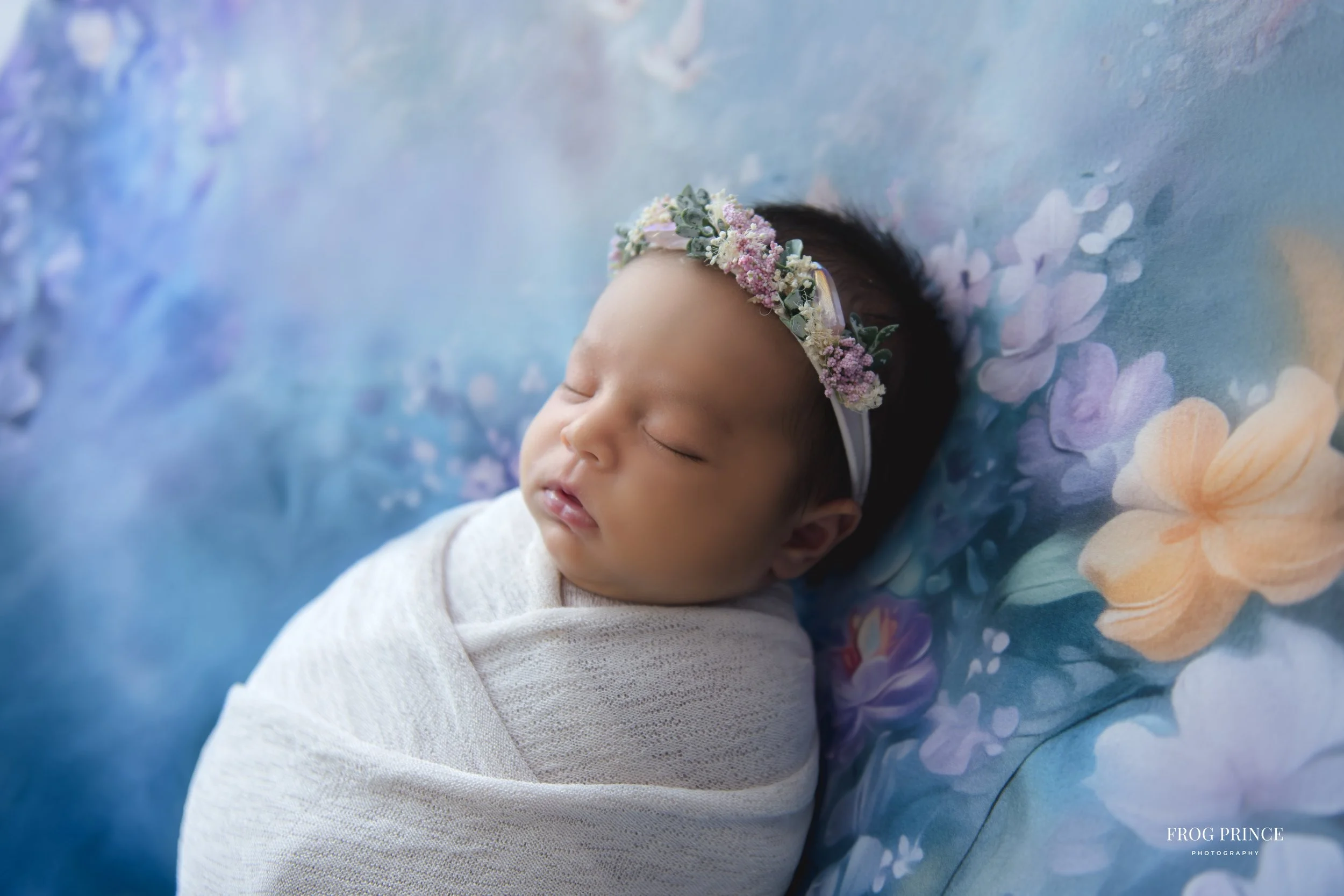 Close-up of a sleeping baby wrapped in a white blanket, wearing a floral headband, lying on a colorful floral background.