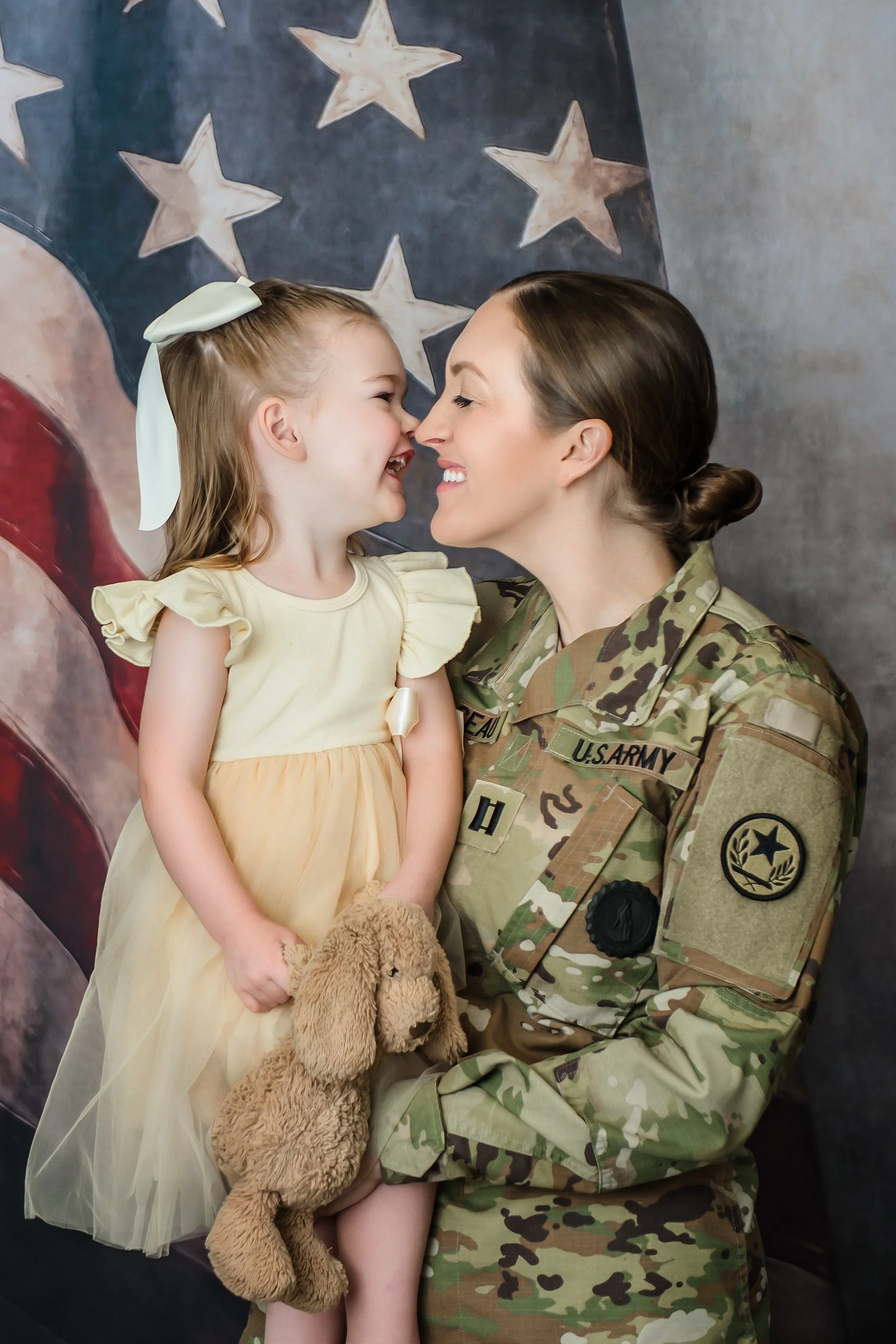 A woman in U.S. Army camouflage uniform holding a young girl in a yellow dress, both touching noses and smiling. The girl holds a stuffed dog. In the background, there is a patriotic American flag.