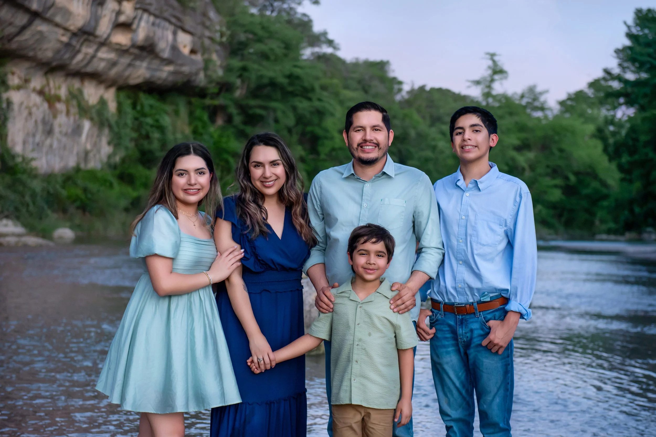 A family of six standing together by a river with green trees and rocky cliffs in the background, smiling at the camera.