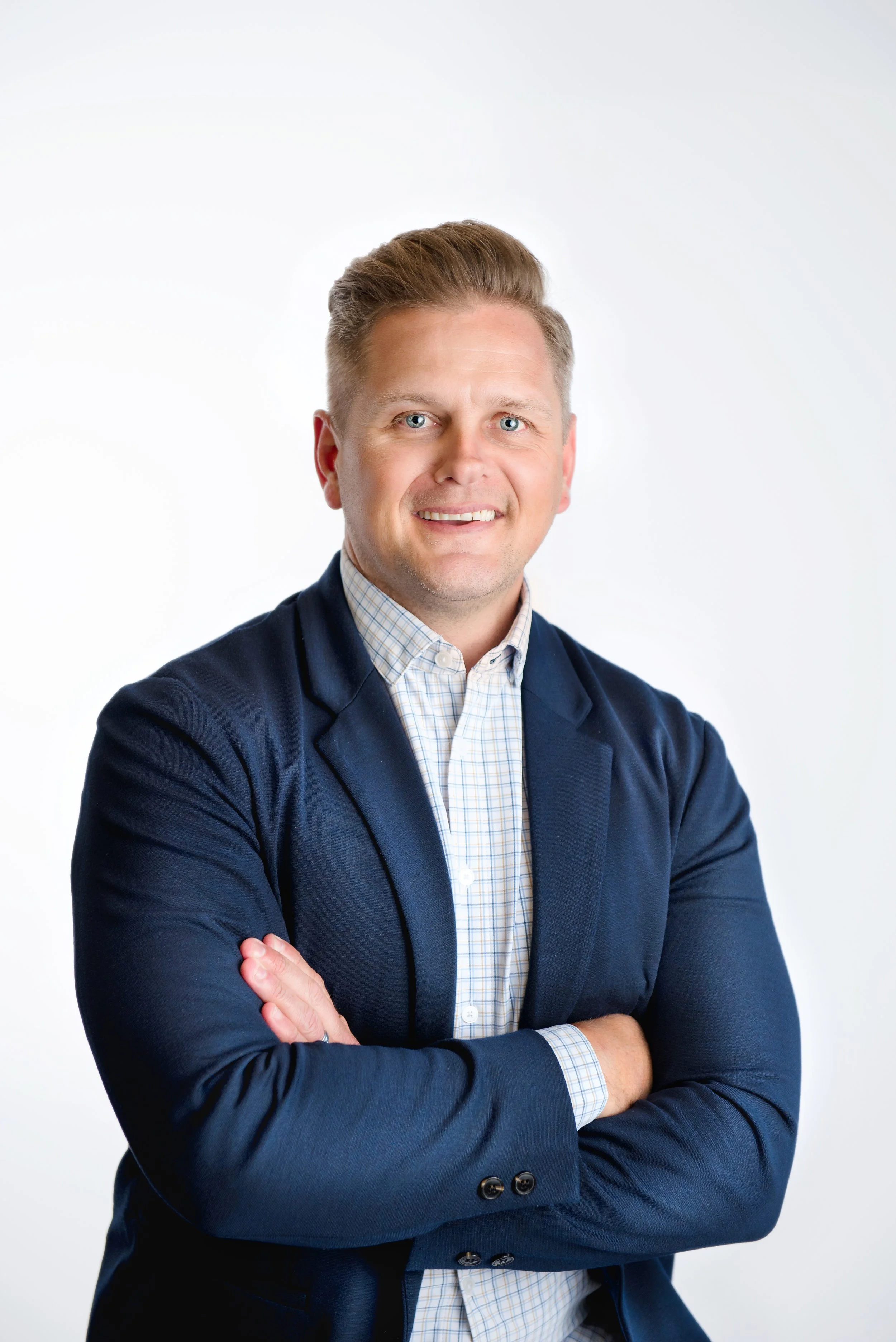Portrait of a smiling man with crossed arms, wearing a suit jacket and a checkered dress shirt, against a plain white background.