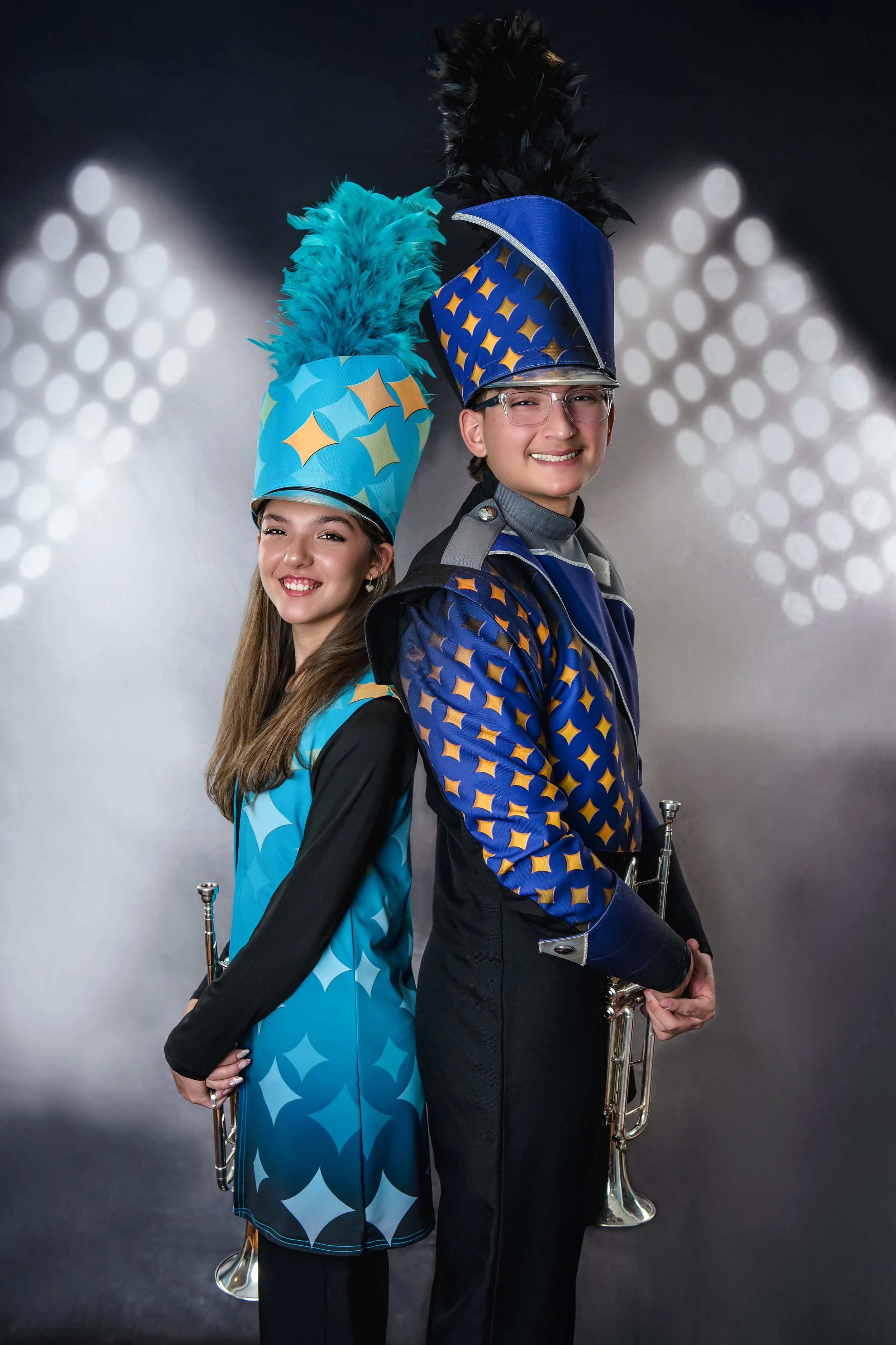 Two young band members, a girl and a boy, in colorful marching band uniforms with tall feathered hats, smiling and posing with their instruments, against a bokeh background of lights.