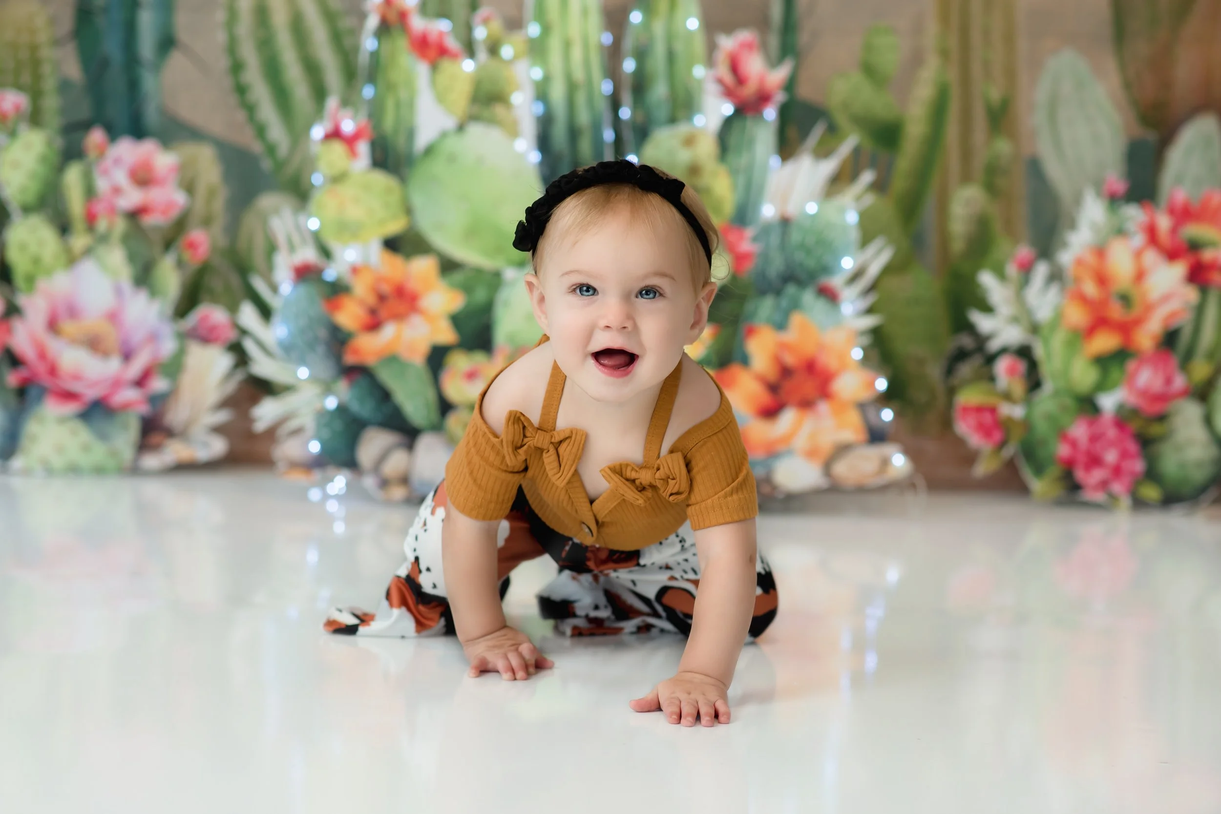 A baby crawling on a white floor, wearing a mustard yellow top with bow details and patterned pants, in front of a colorful botanical background.