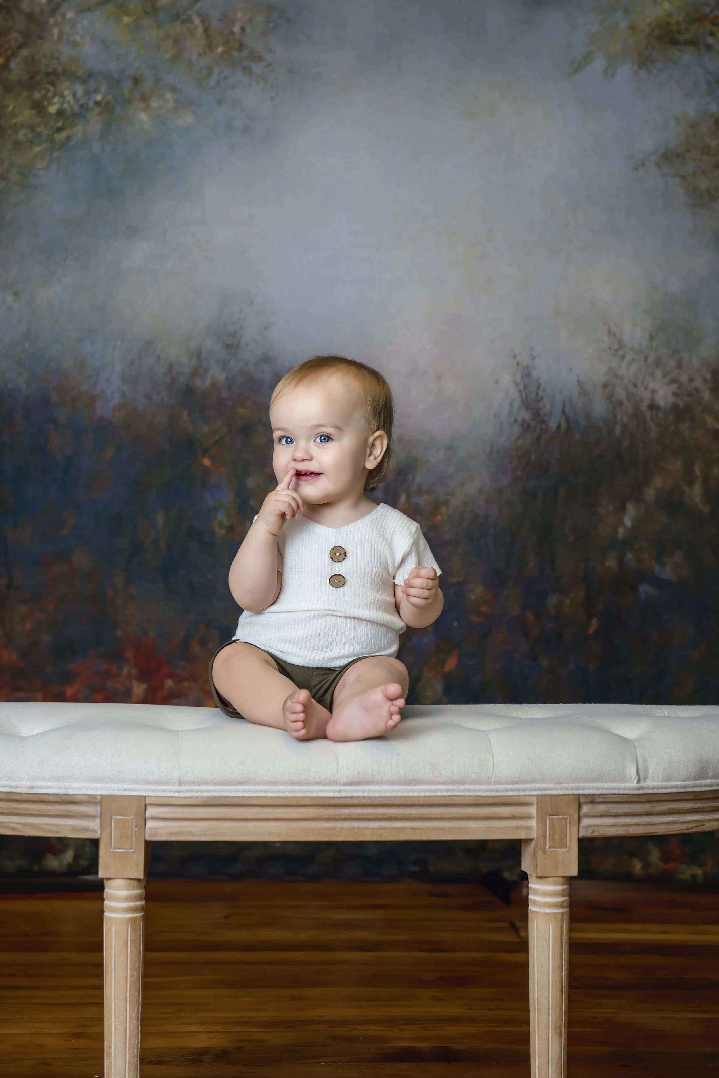 A young toddler sitting on a white cushioned bench with wooden legs, touching their lips with one finger, against a dark, textured background in our Spring Branch studio.