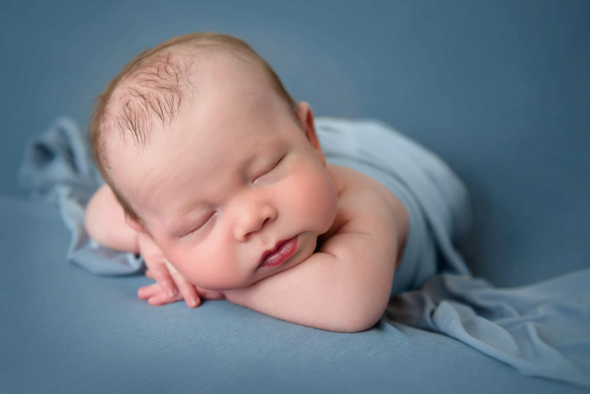 Fine art newborn portrait of a sleeping baby in a light blue outfit resting on a soft blue surface with head gently supported by their hand.