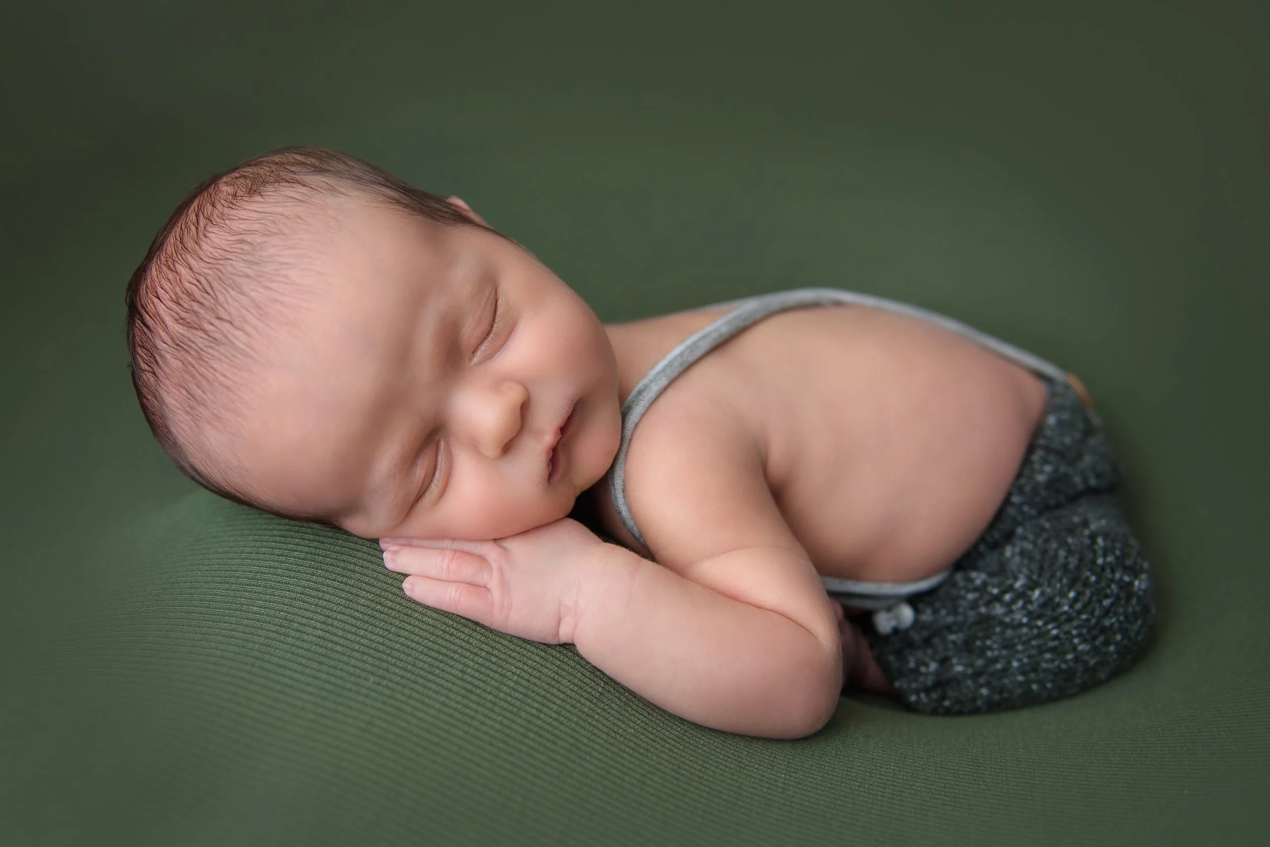Fine art newborn portrait of a sleeping baby in neutral styling posed on a soft green surface with hand tucked under face.