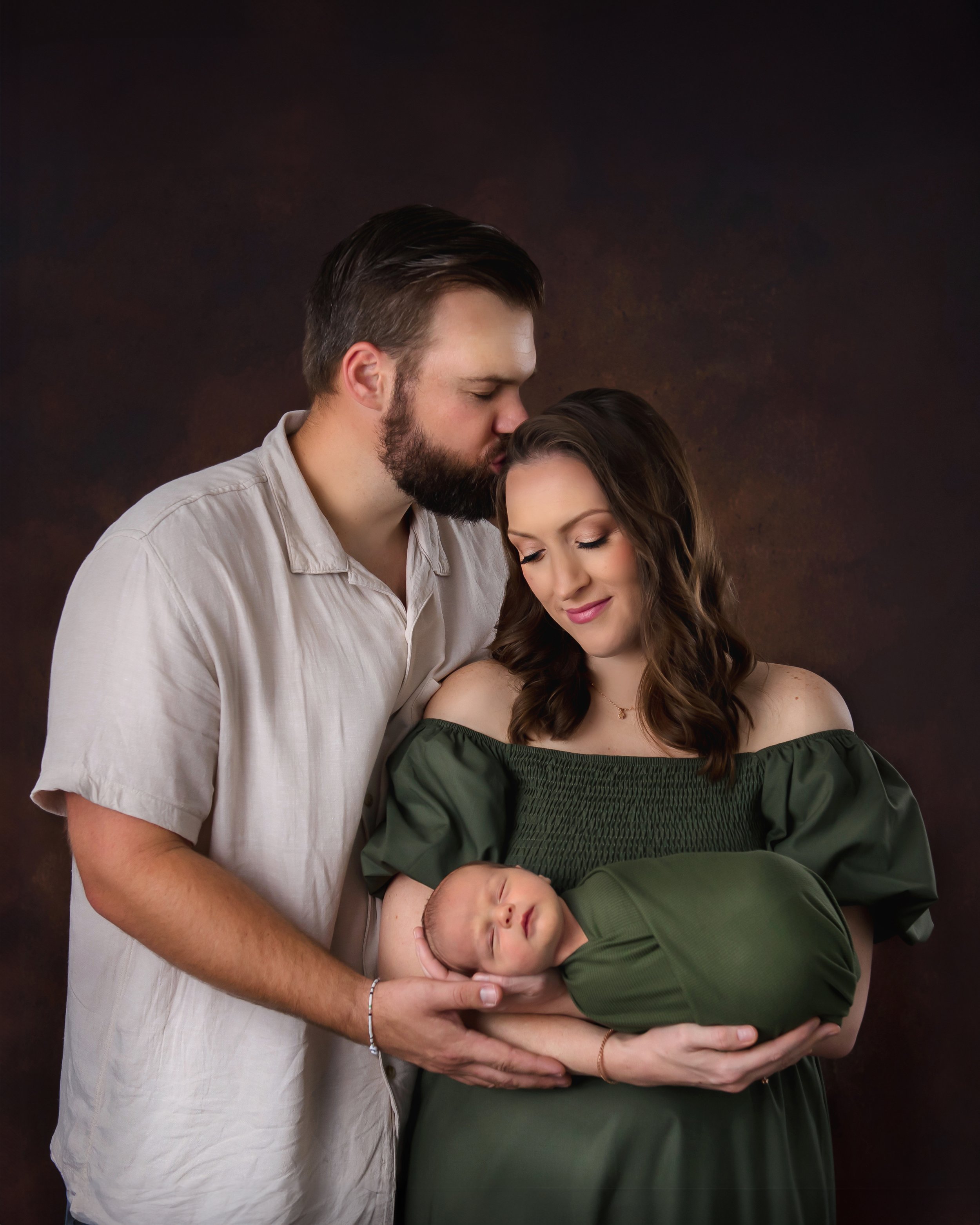 Luxury newborn family portrait of parents holding their sleeping baby in an intimate studio setting with a textured dark backdrop.