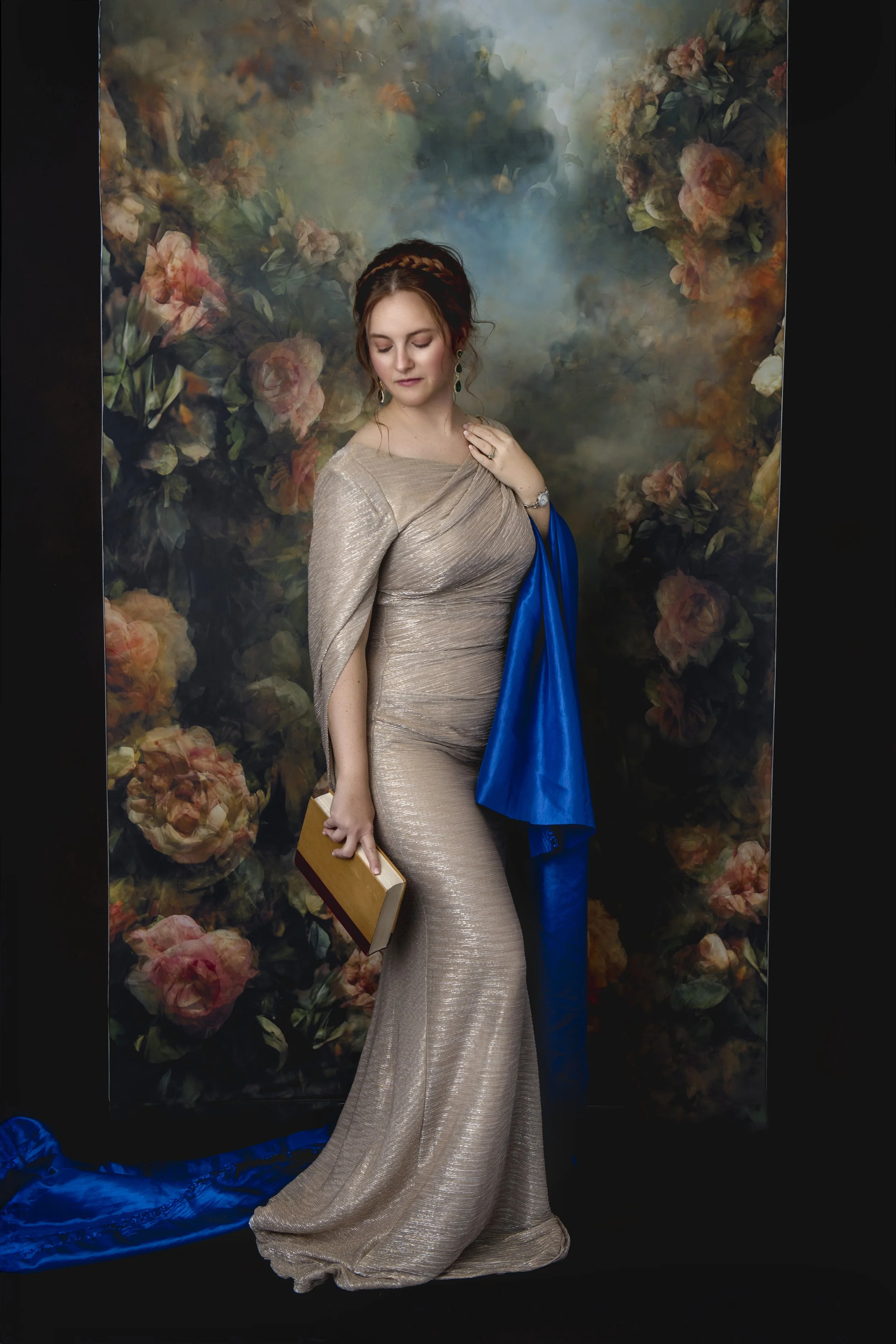 A woman in a long, shimmering beige gown holding a small, brown, hardcover book. She stands against a floral background with pink and peach roses, and a piece of blue fabric lays on the ground.