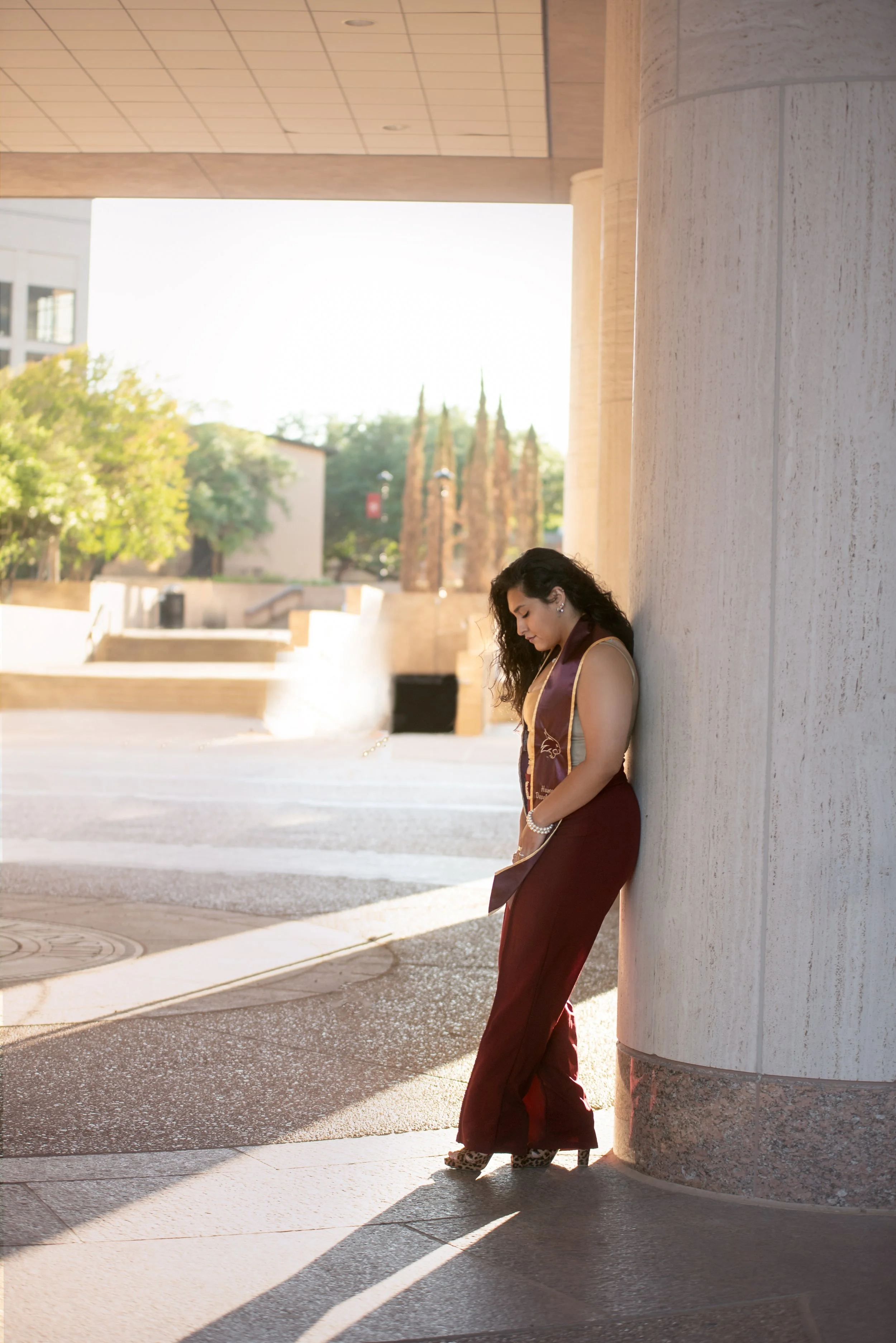 A young woman in a maroon graduation gown and dress, standing against a concrete pillar outdoors during daylight, looking down with her eyes closed.