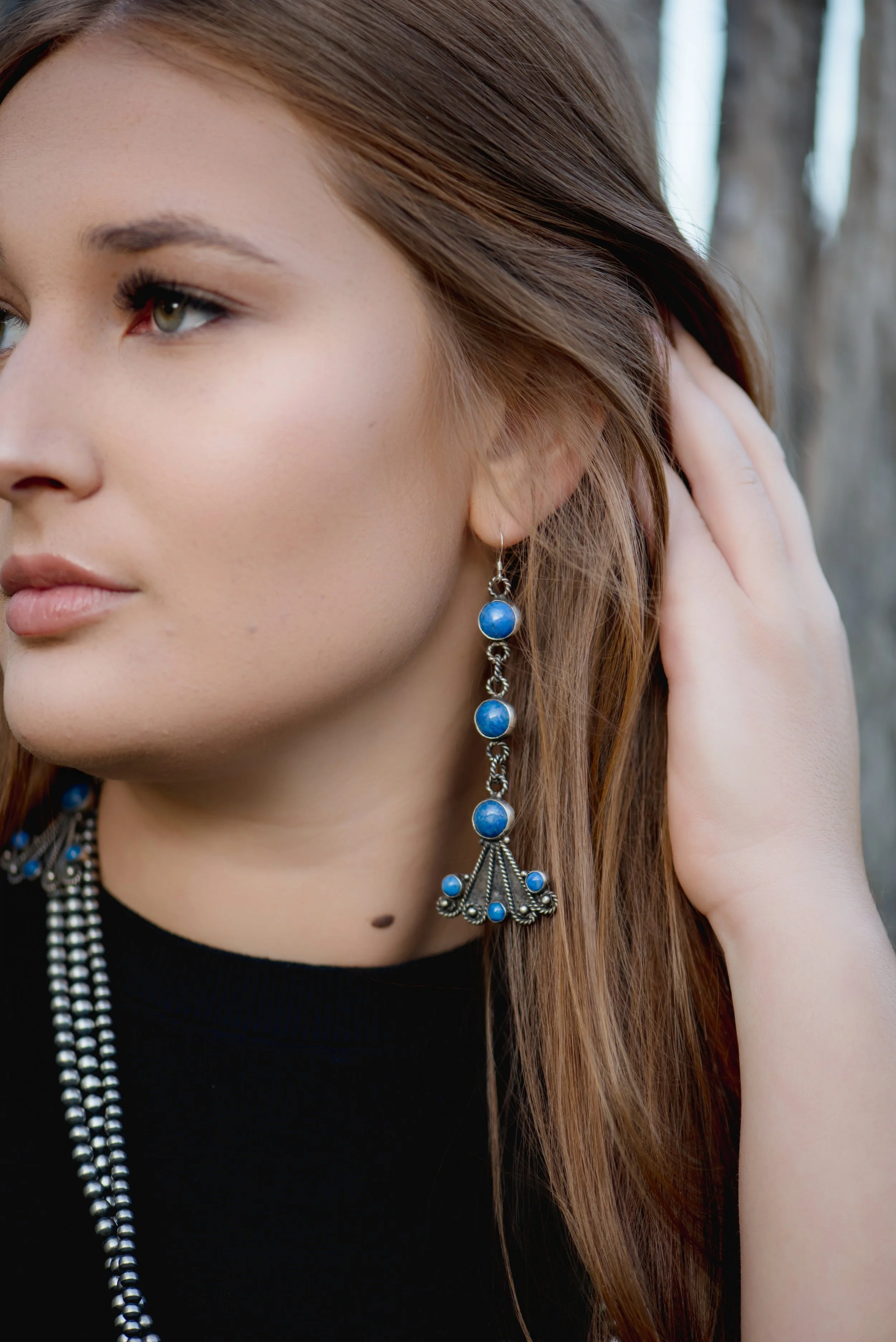 Close-up of a woman with long brown hair wearing blue beaded earrings and a black top, outdoors with trees in the background.