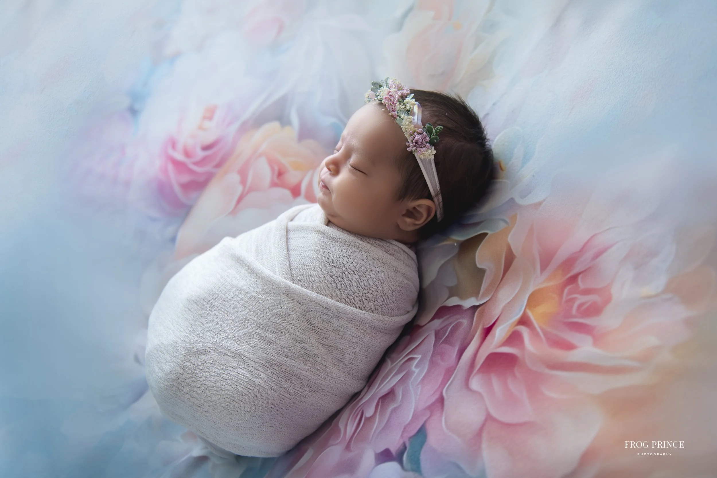 A sleeping baby girl wrapped in a white blanket, lying on a colorful floral blanket with pink, blue, and peach tones, wearing a floral headband.