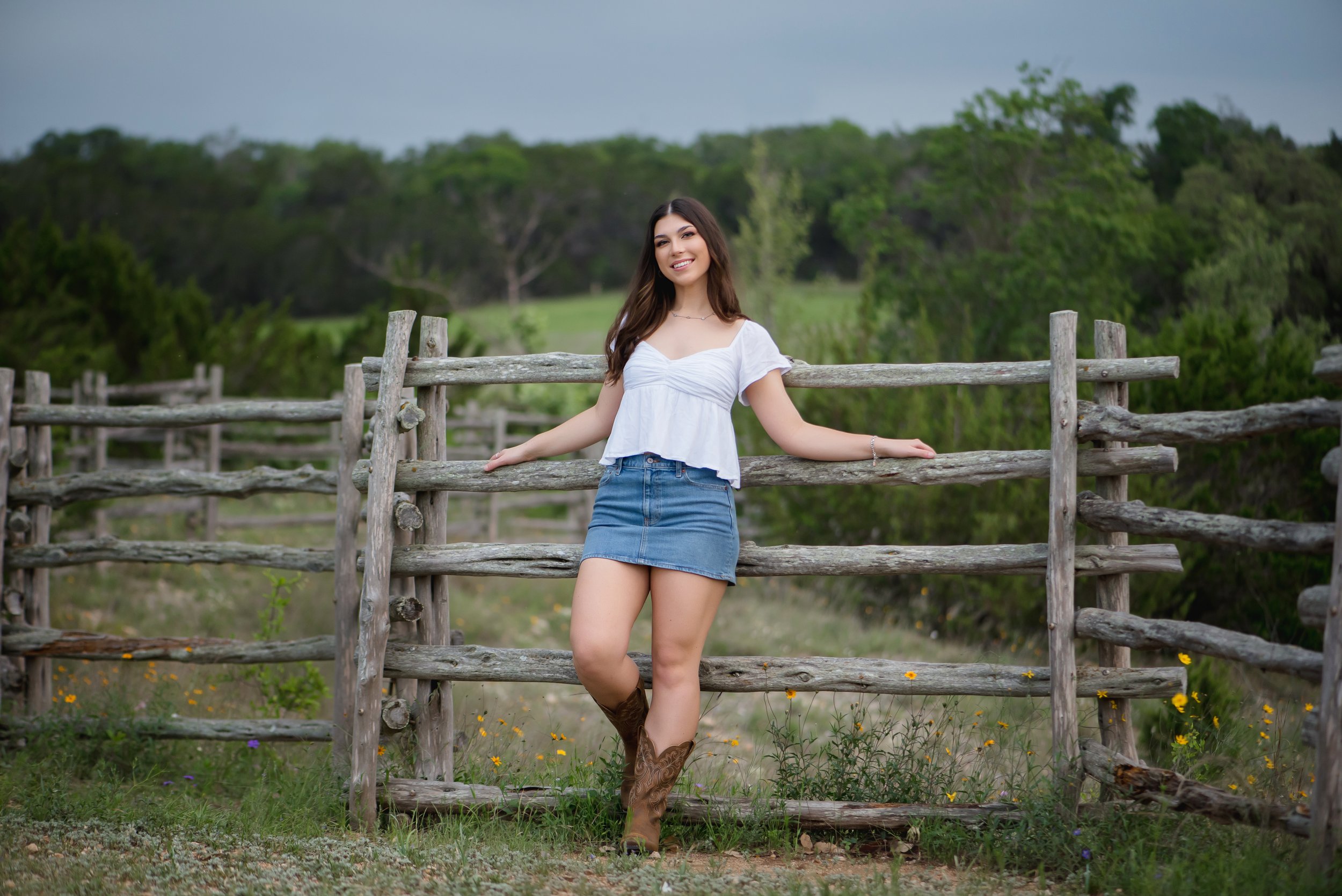 A young woman leaning on a rustic wooden fence in a rural outdoor setting with green trees and grass, smiling at the camera.
