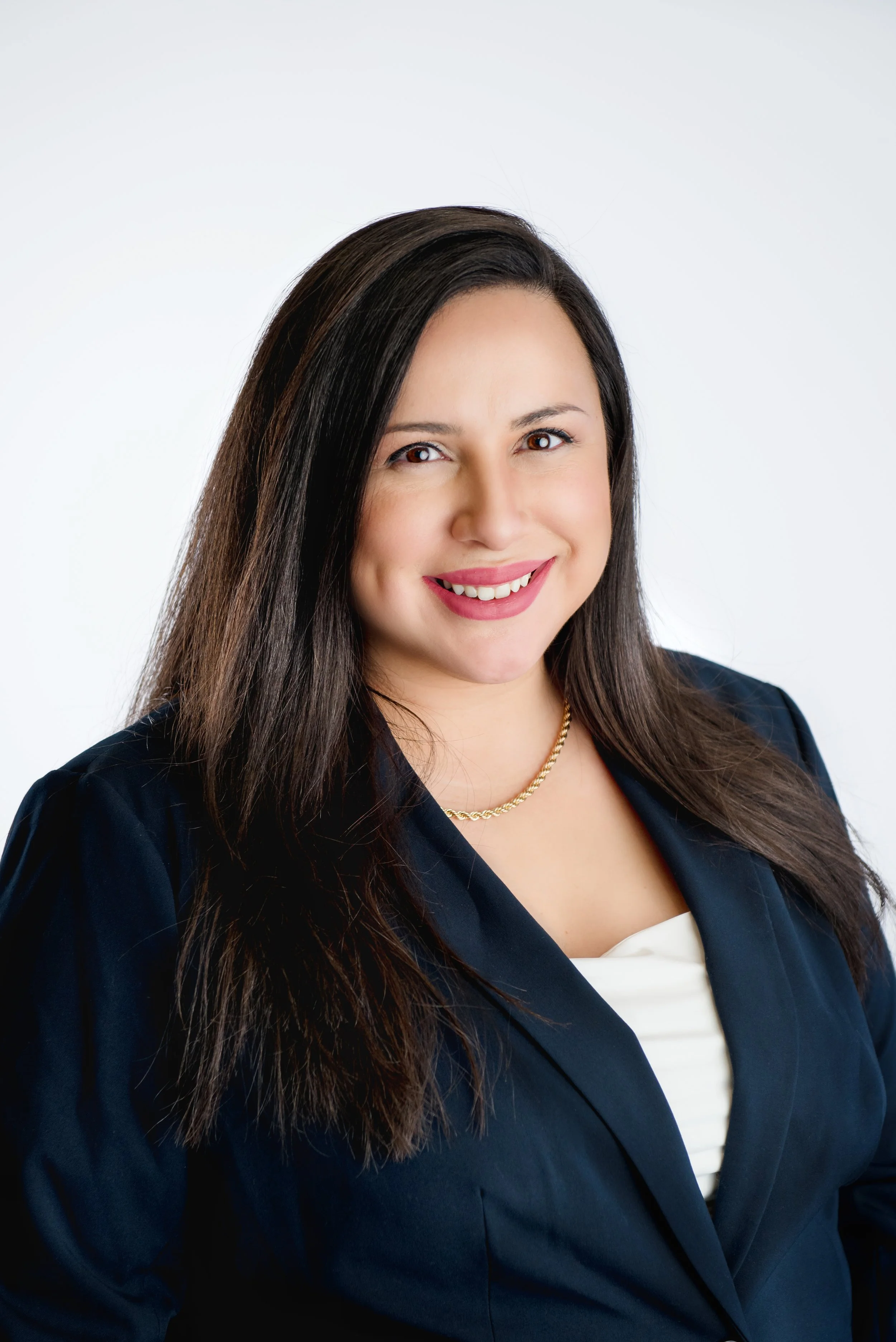 A professional woman with long dark hair smiling at the camera, wearing a dark blazer, a white top, and a gold chain necklace, against a white background.