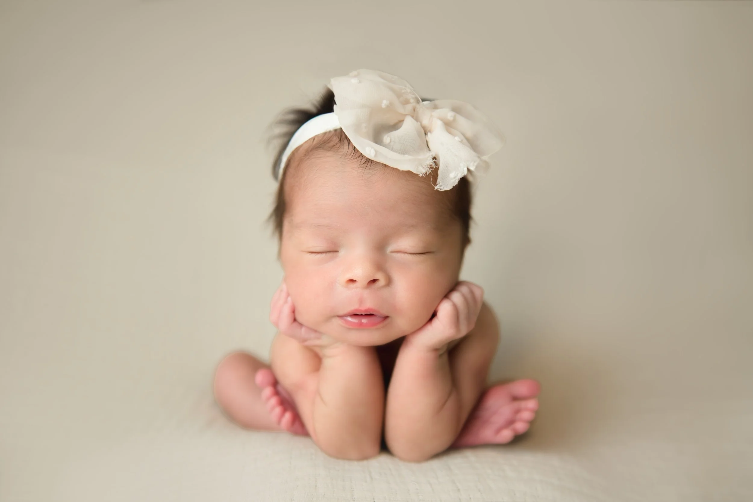 A newborn baby with a large cream-colored bow headband, resting on their hands with eyes closed on a beige surface.