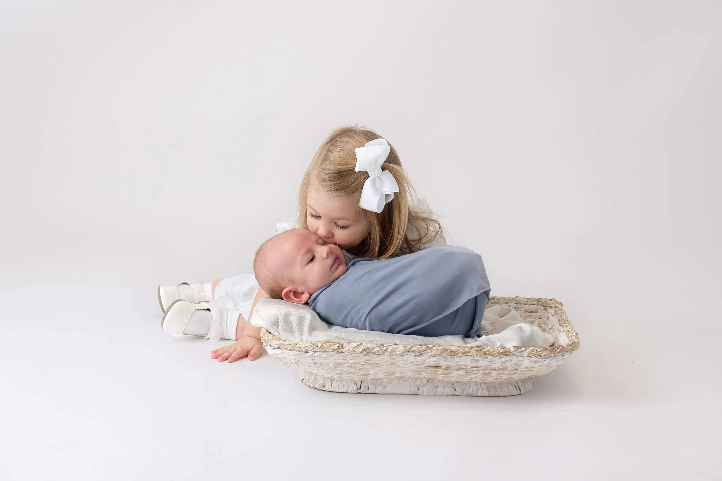 A young girl giving a gentle kiss to a baby wrapped in a blue blanket, lying on a woven basket, against a plain white background.