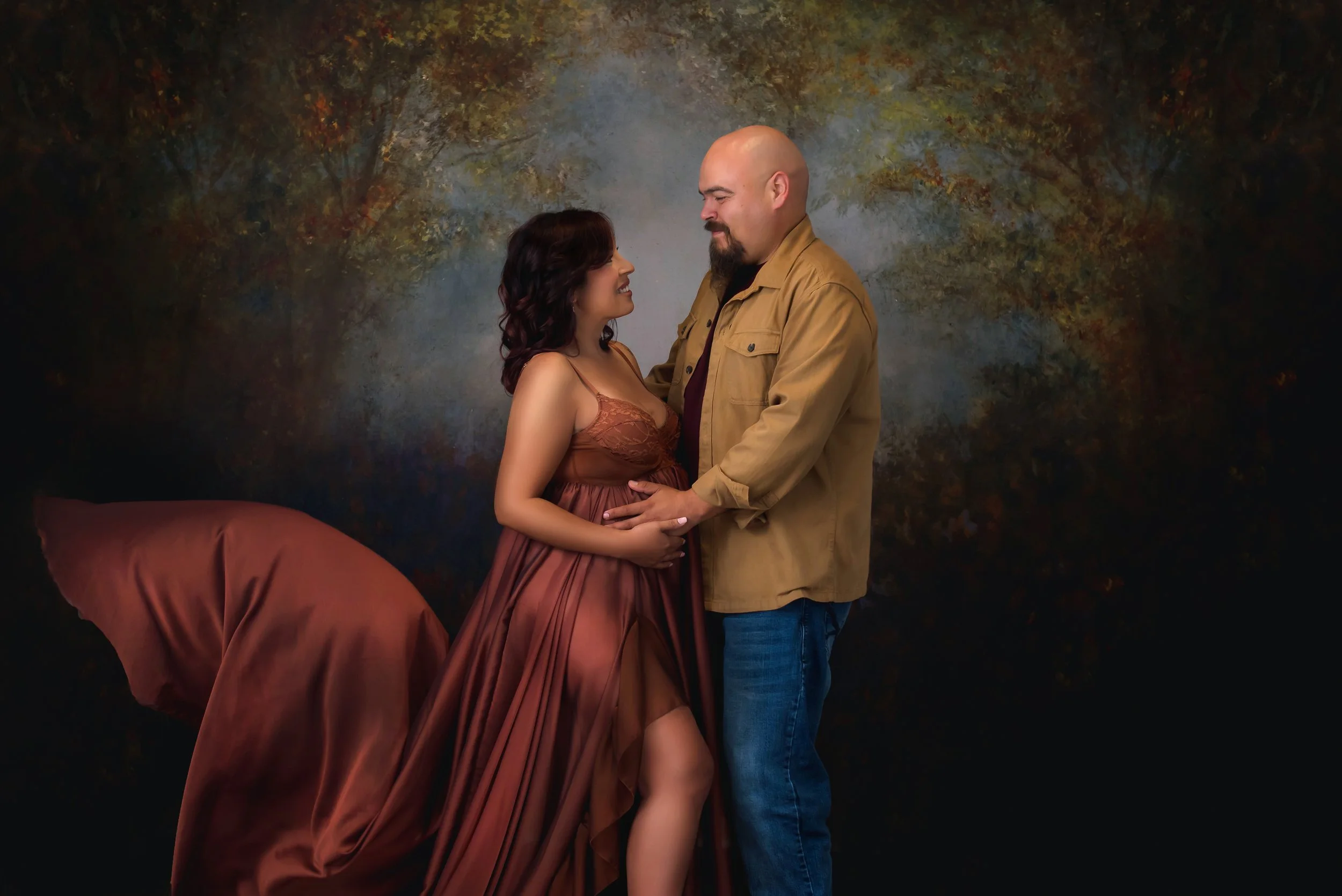 Editorial maternity portrait of a couple standing close together in a romantic embrace, styled in warm neutral tones against a dramatic textured studio backdrop.
