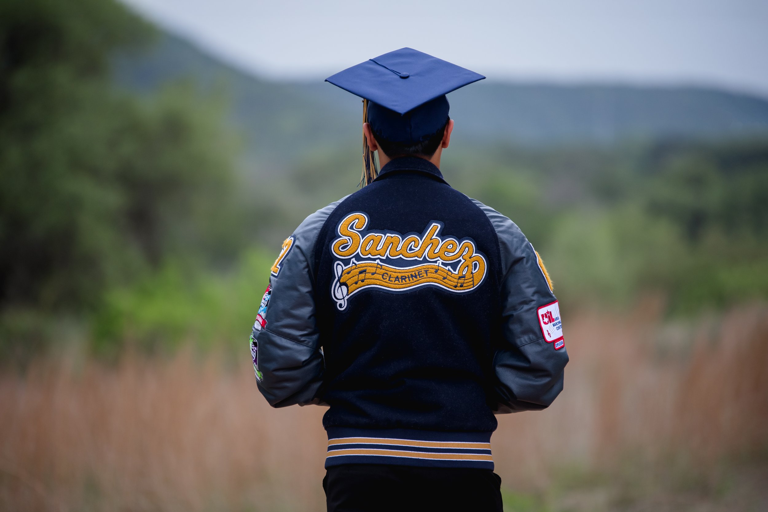 Person wearing a Varsity jacket and graduation cap, standing outdoors with mountains and greenery in the background.