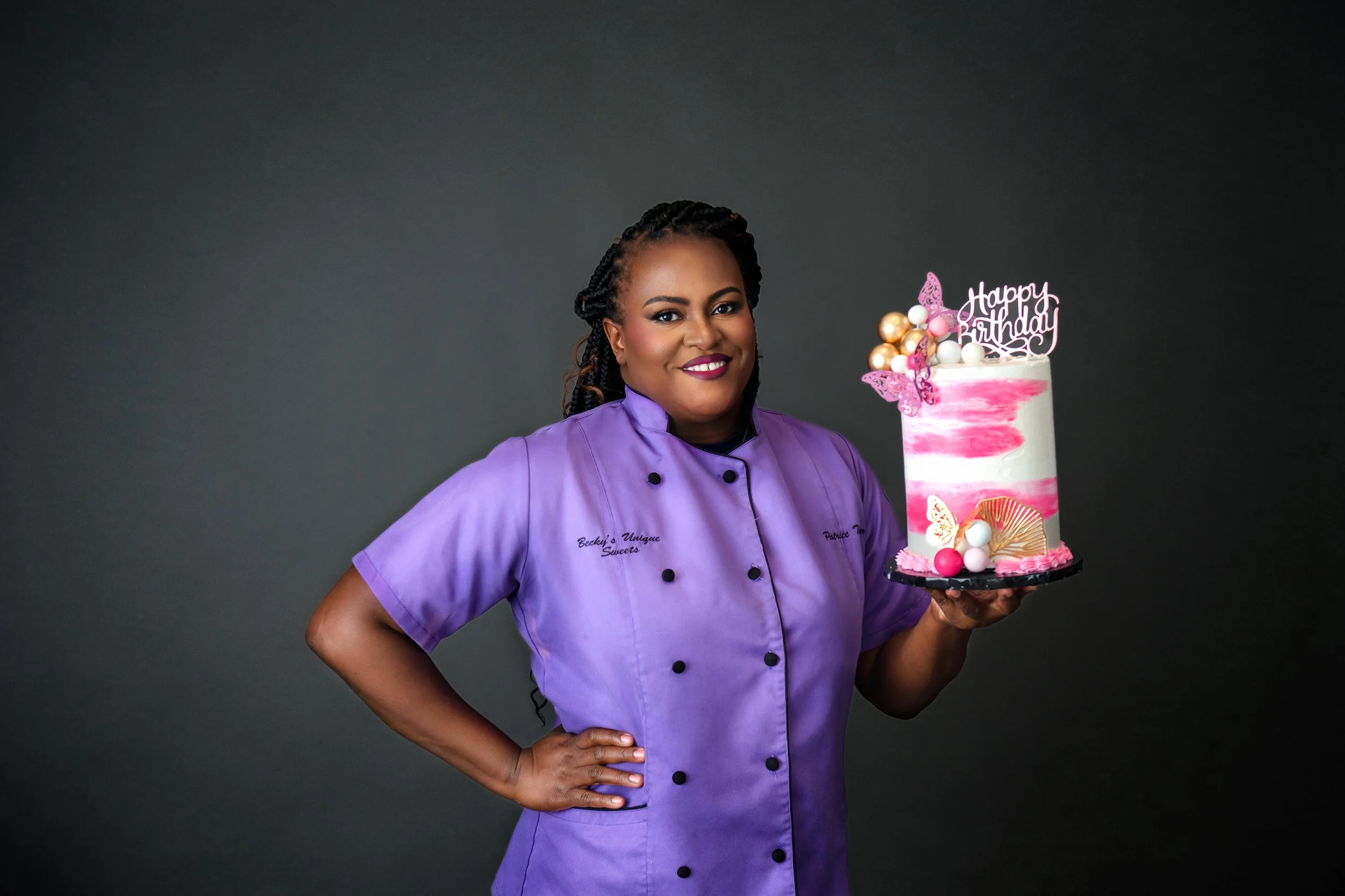 A woman in a purple chef's coat holding a birthday cake with pink, white, and gold decorations against a dark background.