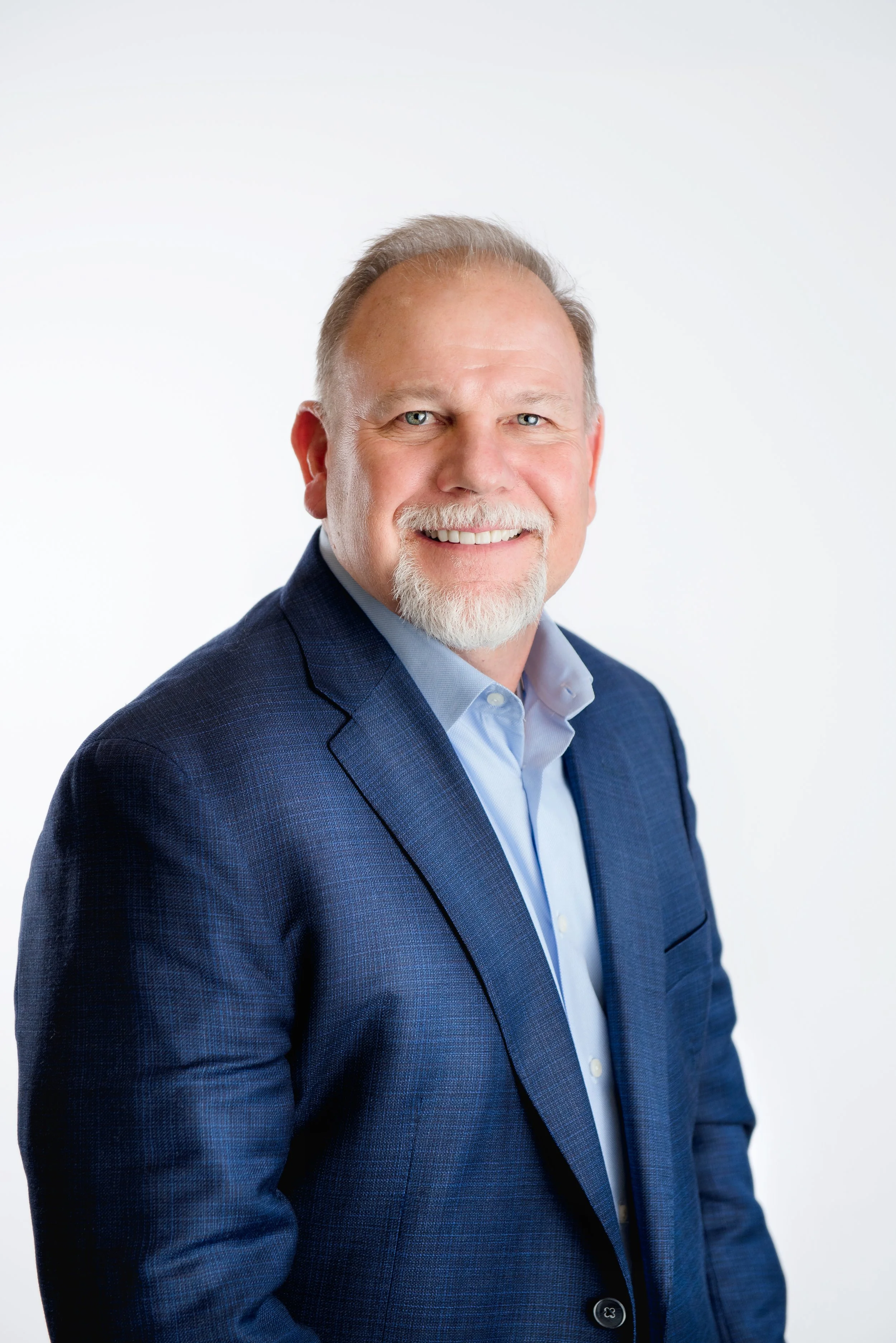 Headshot of a middle-aged man with a beard, wearing a blue suit and light blue shirt, smiling in front of a plain white background.